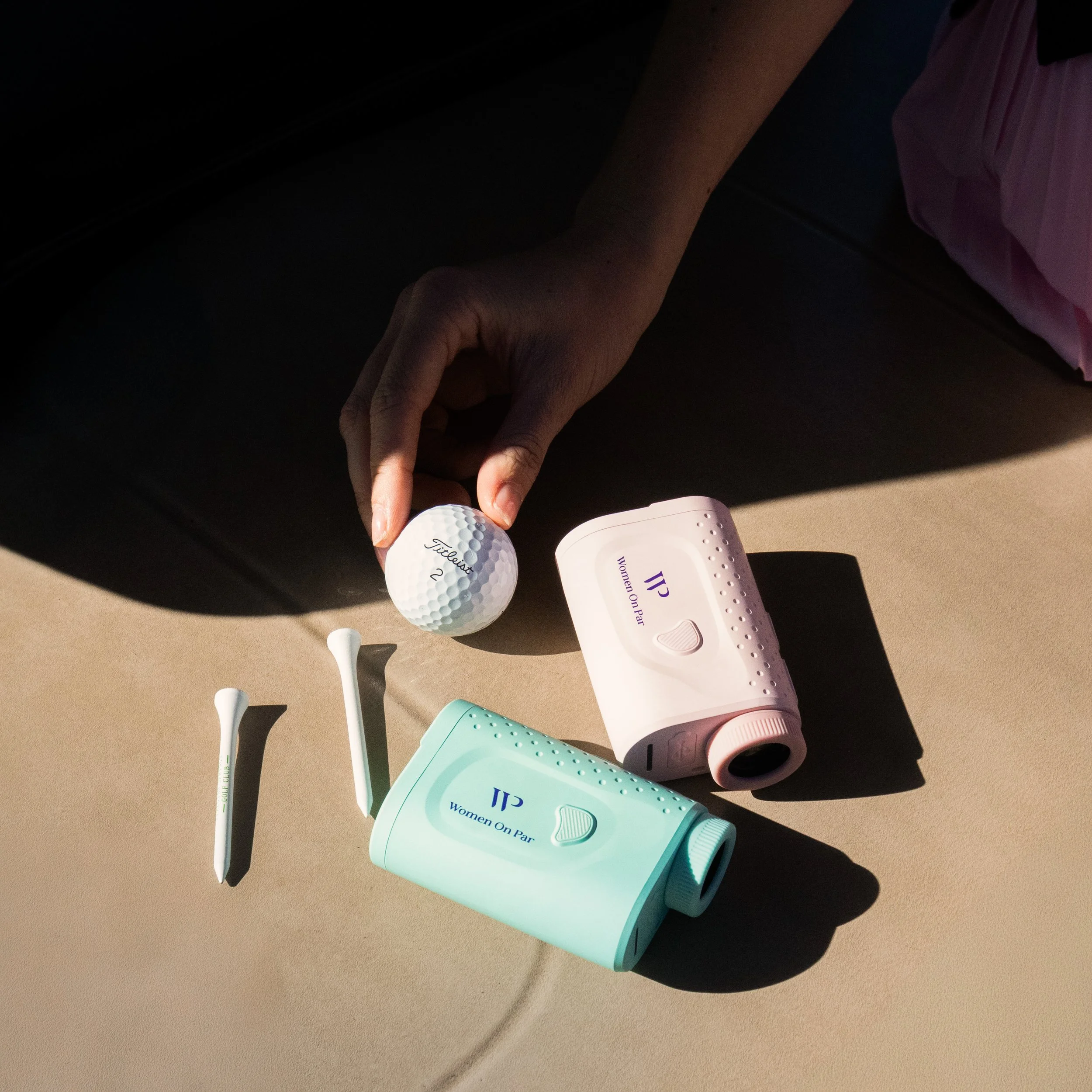 Close-up of a person's hand holding a golf ball marked with 'Titleist 2' next to a pink medical or skincare device labeled 'Women on Par,' with golf clubs lying on a beige surface.