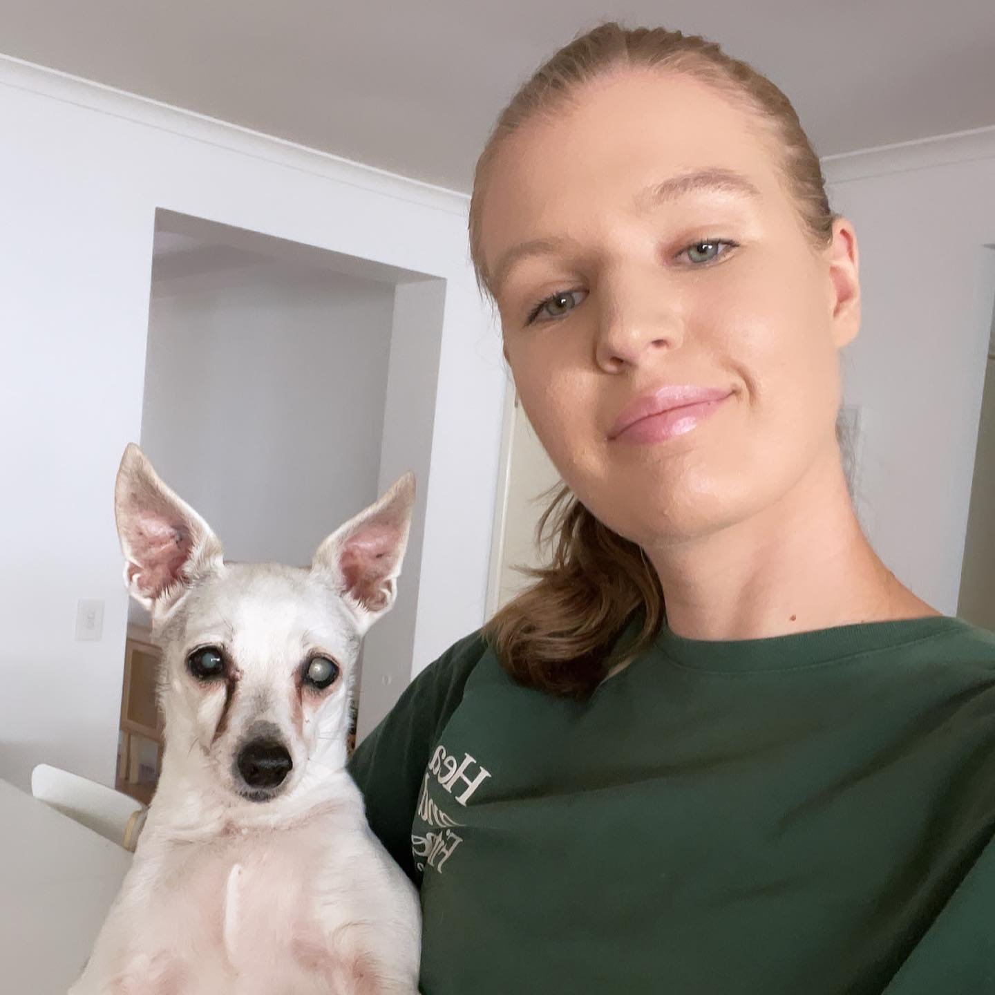 A woman with blonde hair in a ponytail takes a selfie with a white and gray dog inside a home. The woman is smiling and wearing a green shirt, and the dog has upright ears and appears alert.