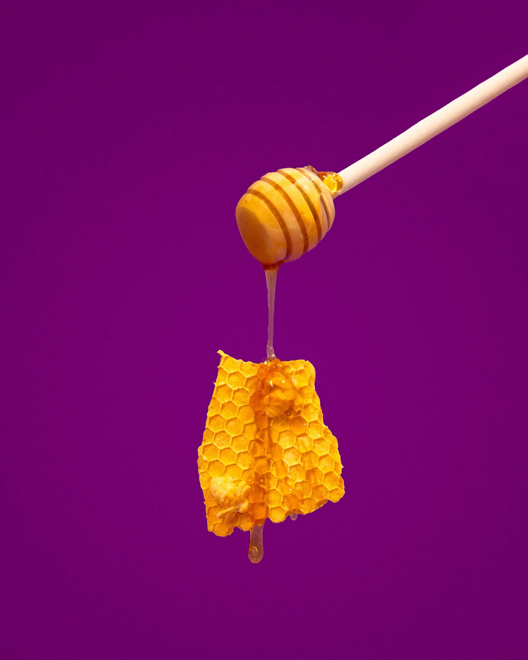 A honey dipper with honey dripping onto a honeycomb against a purple background.