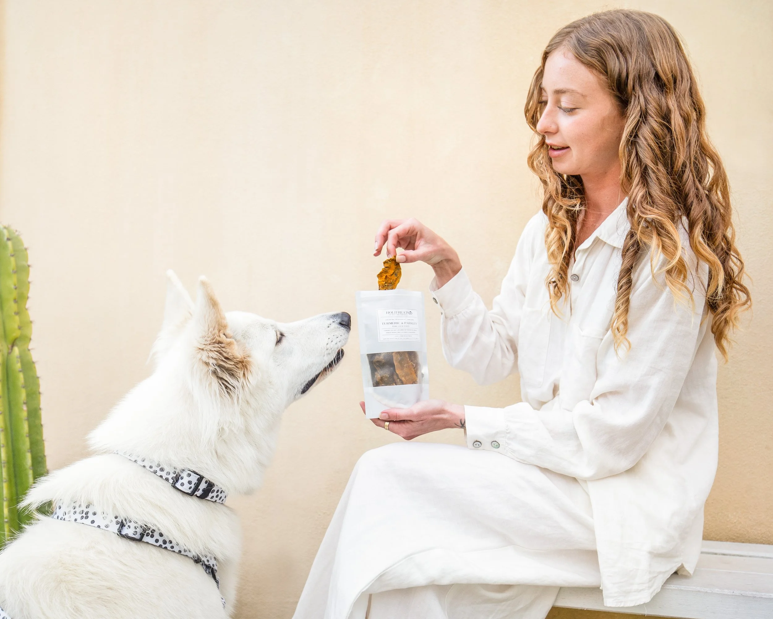 A woman with long, wavy, light brown hair wearing a white shirt, smiling and sitting indoors with a white dog wearing a black and white polka-dotted bandana around its neck. The woman is holding a package of dog treats labeled 'Turmeric & Parsley' an