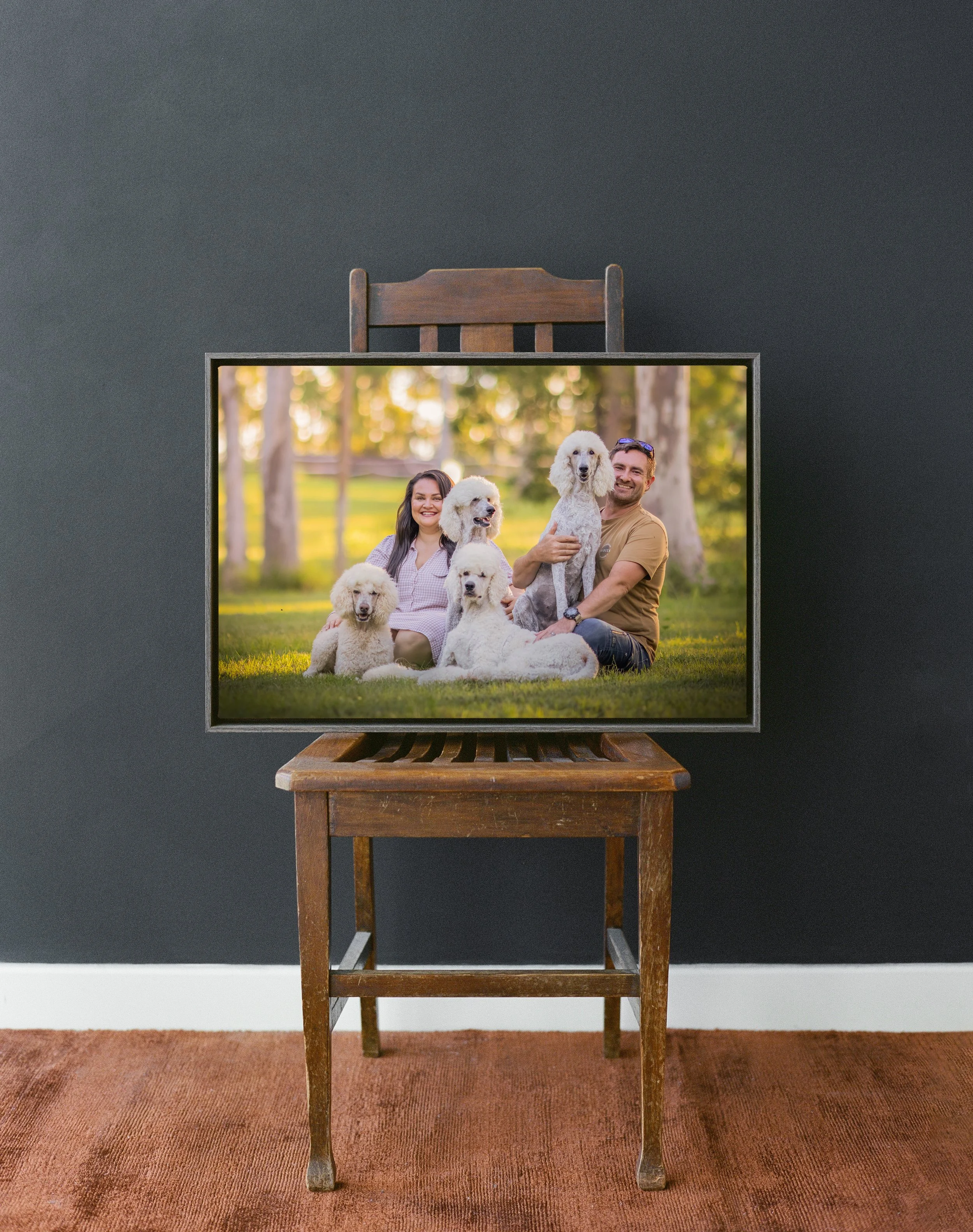 A wooden table with an old, dark chair behind it, set against a dark wall, with a framed photo of a happy couple and their poodles on a grassy park, placed on the table and in front of the chair.