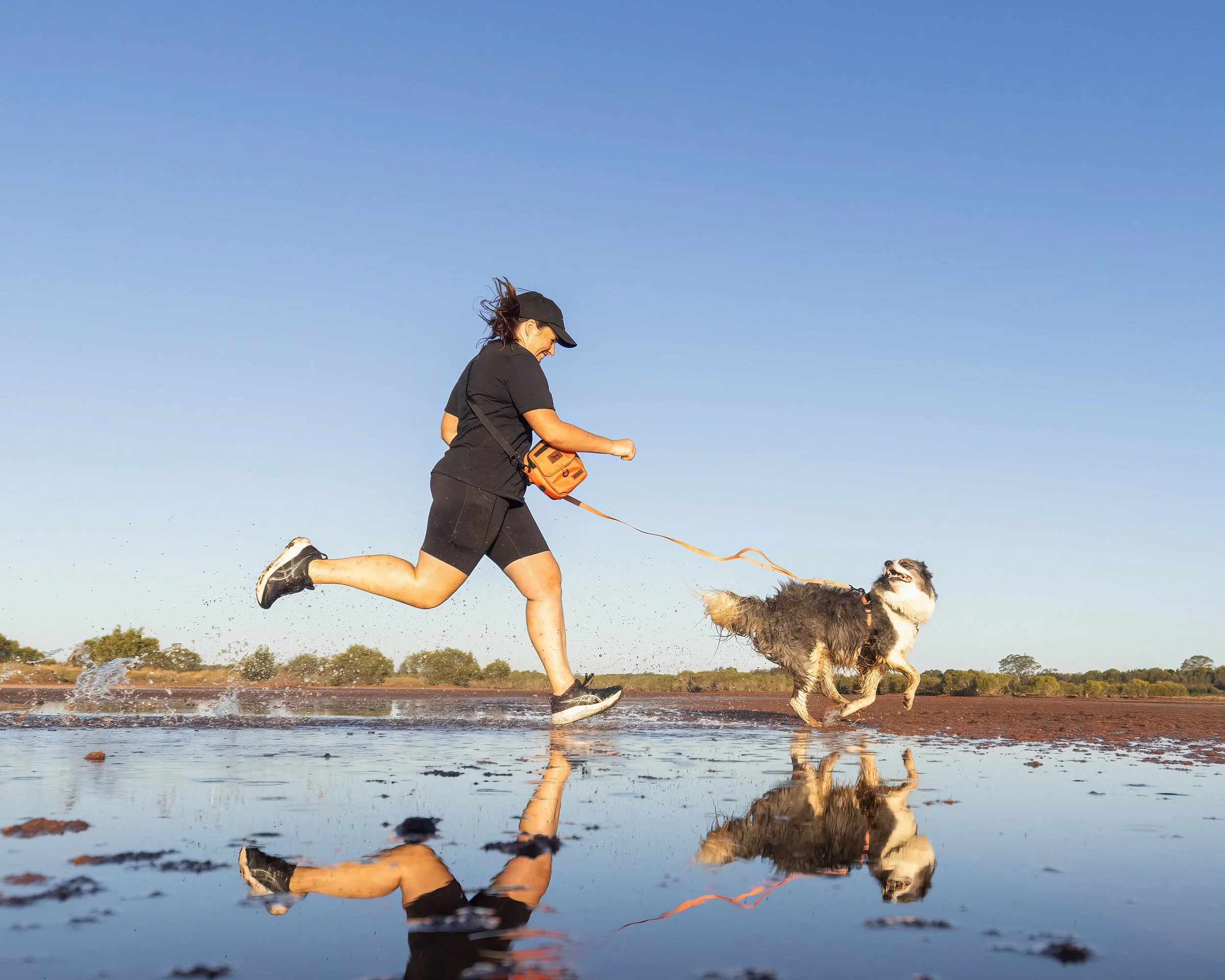 A woman running with a dog through a shallow body of water on a clear day, with trees in the background and a bright blue sky.