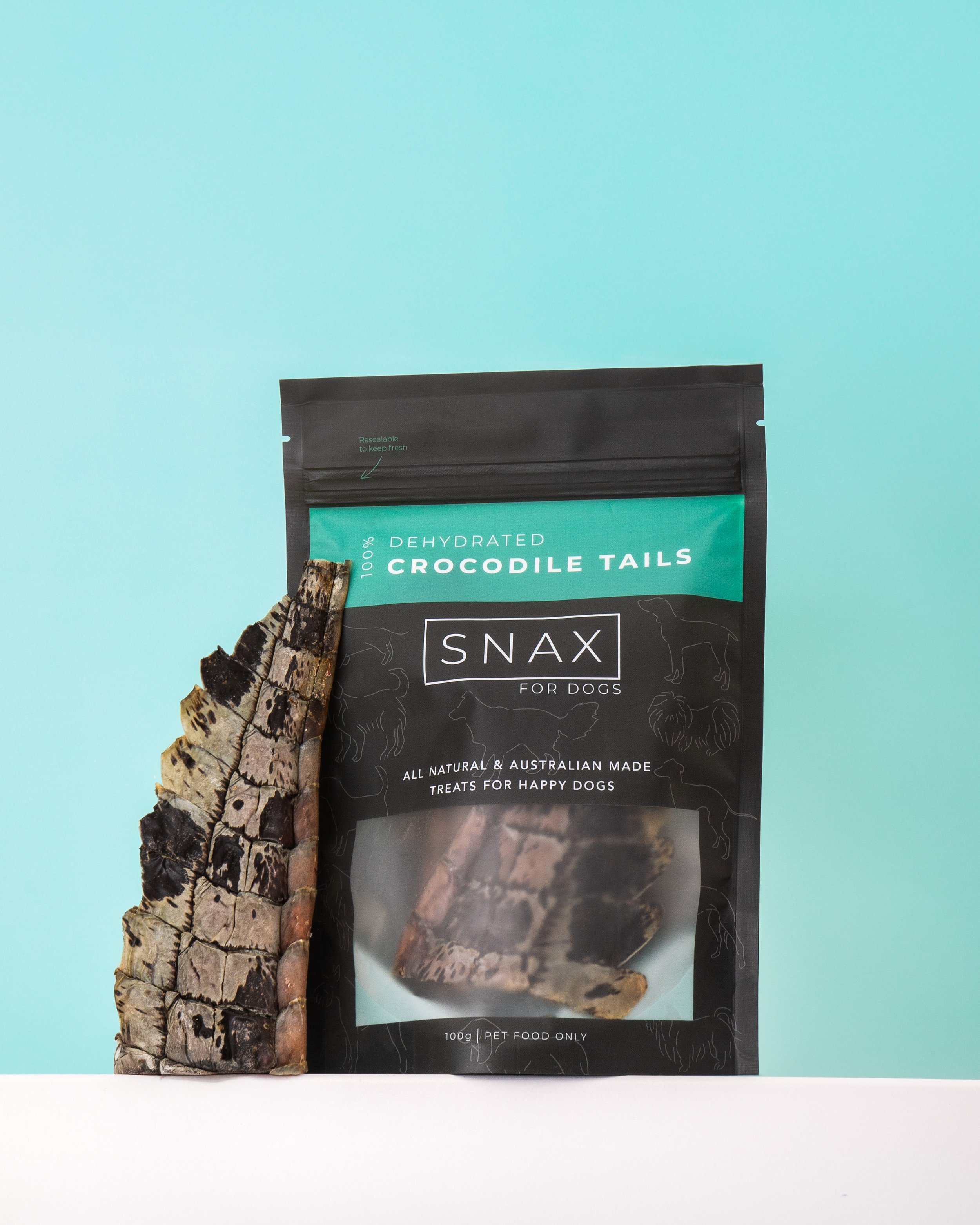 Packaging of dehydrated crocodile tails treats with a piece of crocodile tail attached to the package, against a light blue background.