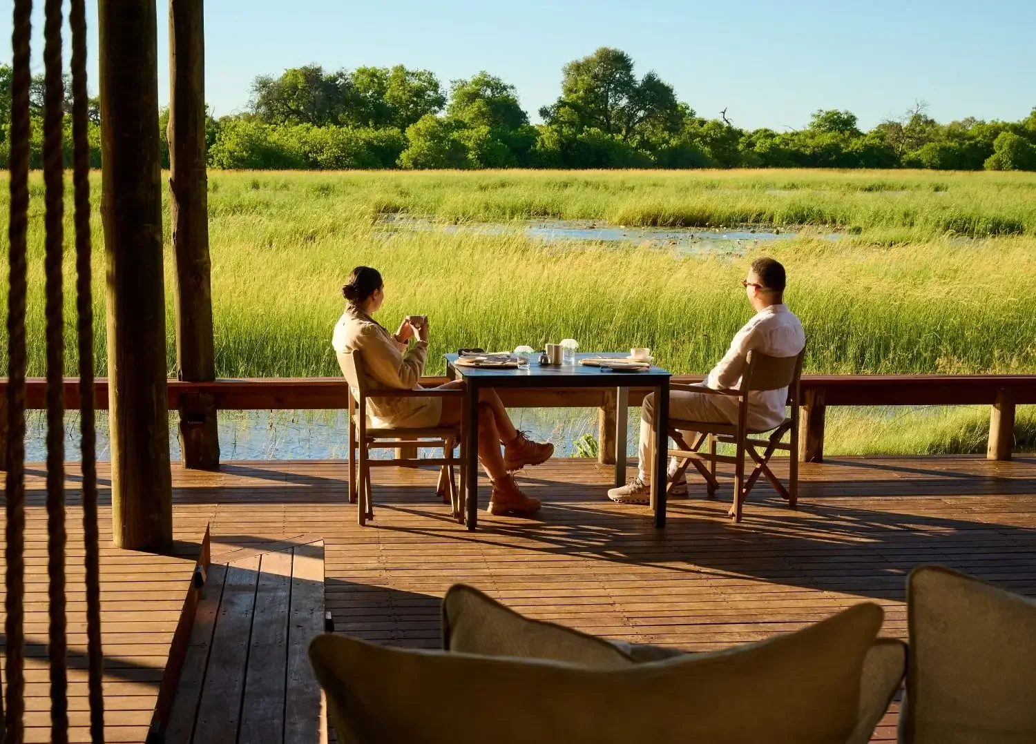 A couple enjoying a private, romantic morning coffee at a table on a wooden deck overlooking the lush green wetlands during a luxury Botswana honeymoon safari.