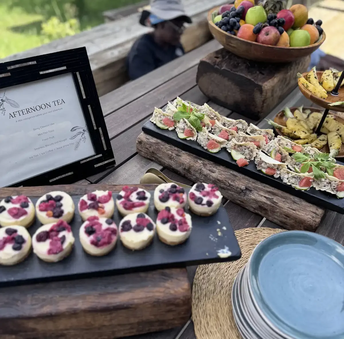 A beautifully arranged afternoon tea spread at a luxury safari camp, featuring artisanal berry cheesecakes, savory snacks, and fresh fruit bowls on a rustic wooden table.
