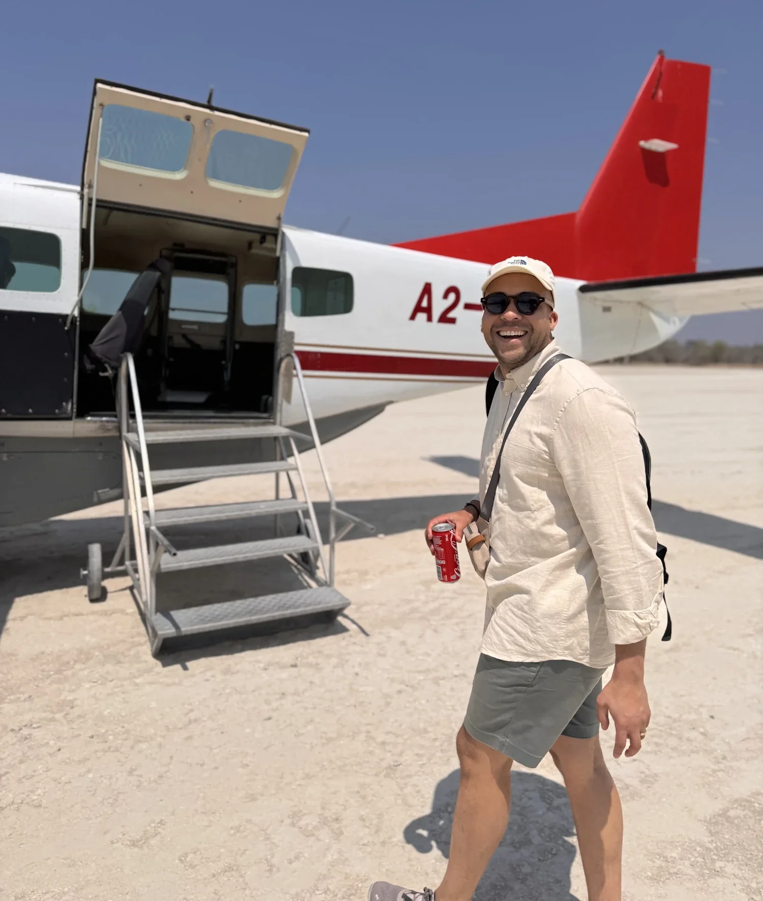 Co-founder James Briscoe wearing sunglasses and smiling while walking away from a white and red bush plane on a sunlit, flat landscape.