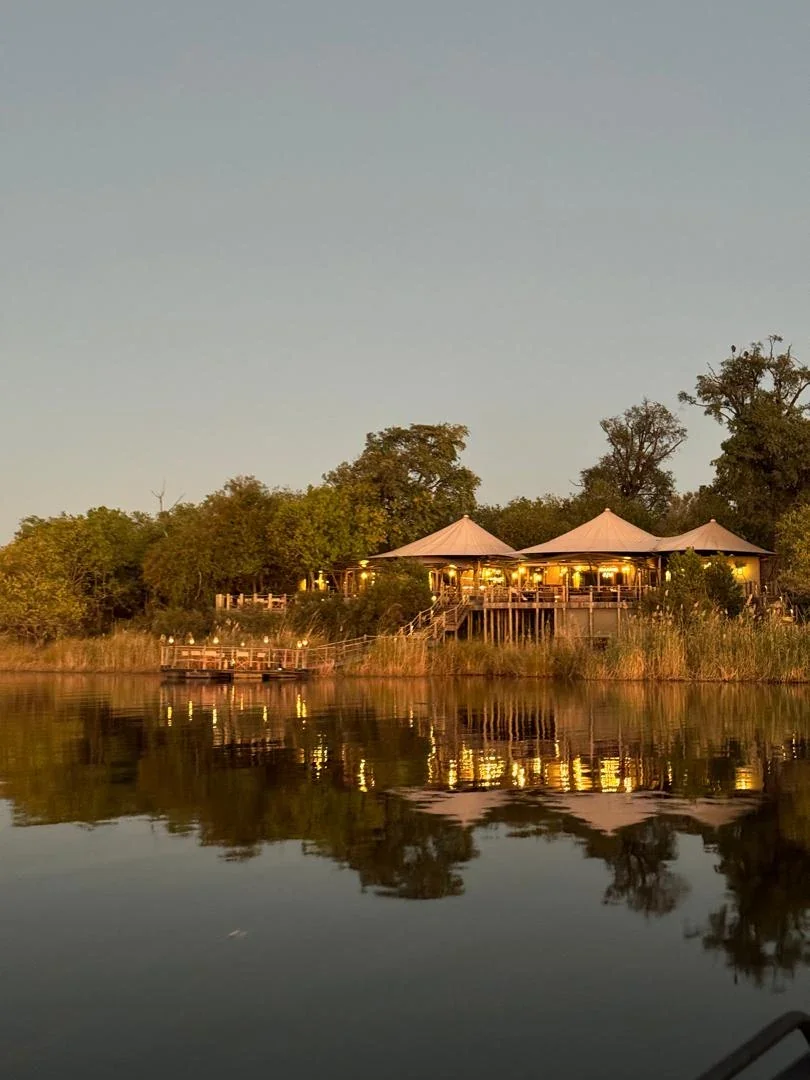 A beautiful luxury tented safari lodge glowing warmly at dusk reflecting in a calm African river