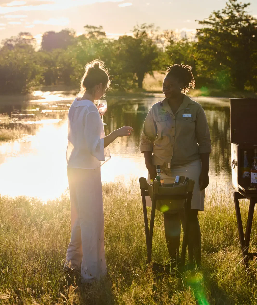 A safari guest interacts warmly with a local hospitality staff member while enjoying drinks in a field during the soft, warm golden hour light in Africa.