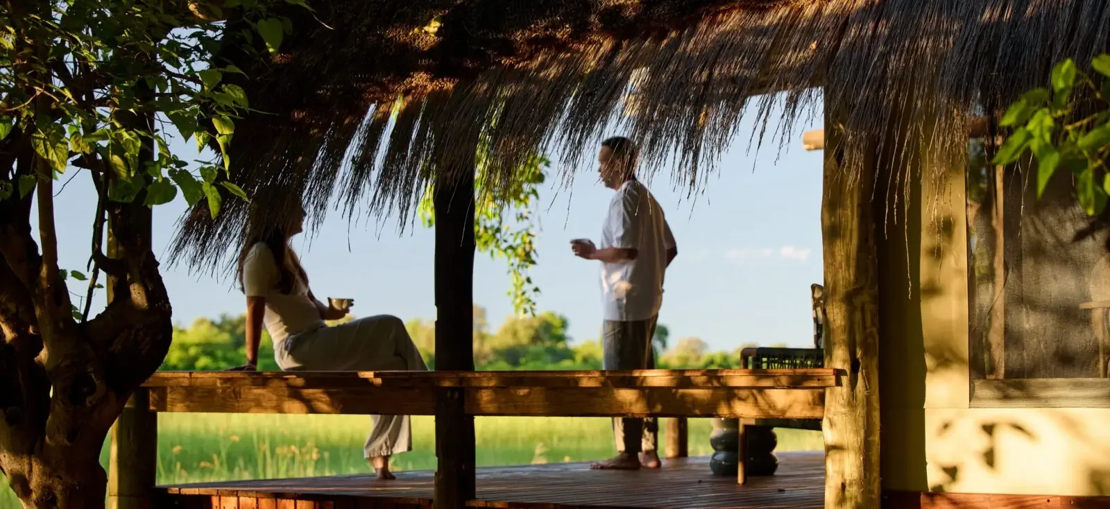 A couple relaxing with drinks on the shaded wooden deck of a luxury safari lodge, looking out over the green Botswana wilderness during a romantic honeymoon getaway.