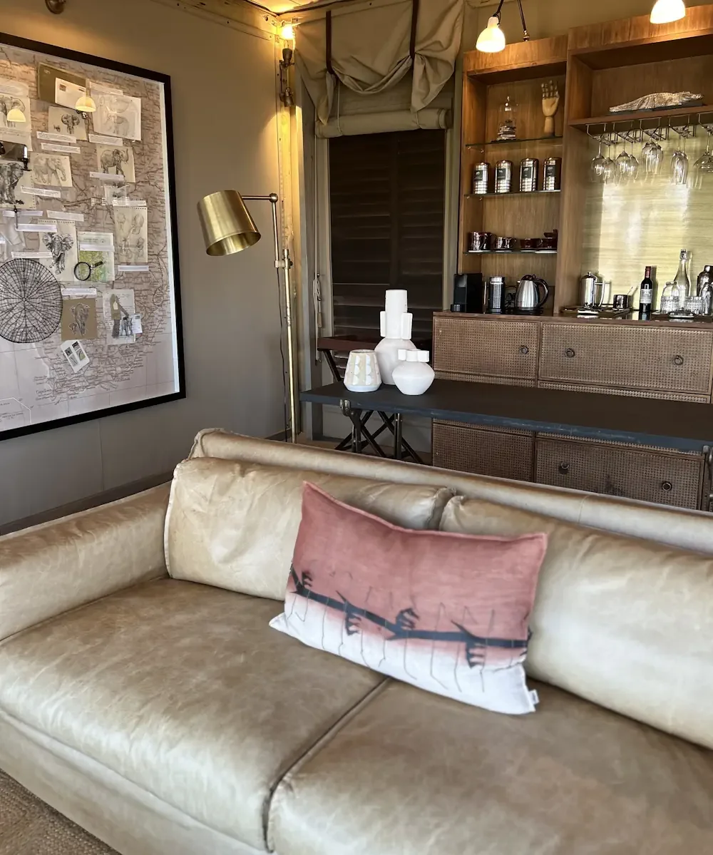 A beautifully appointed luxury safari lodge lounge featuring a plush leather sofa, a well-stocked bar area, and a large vintage map of Africa on the wall.