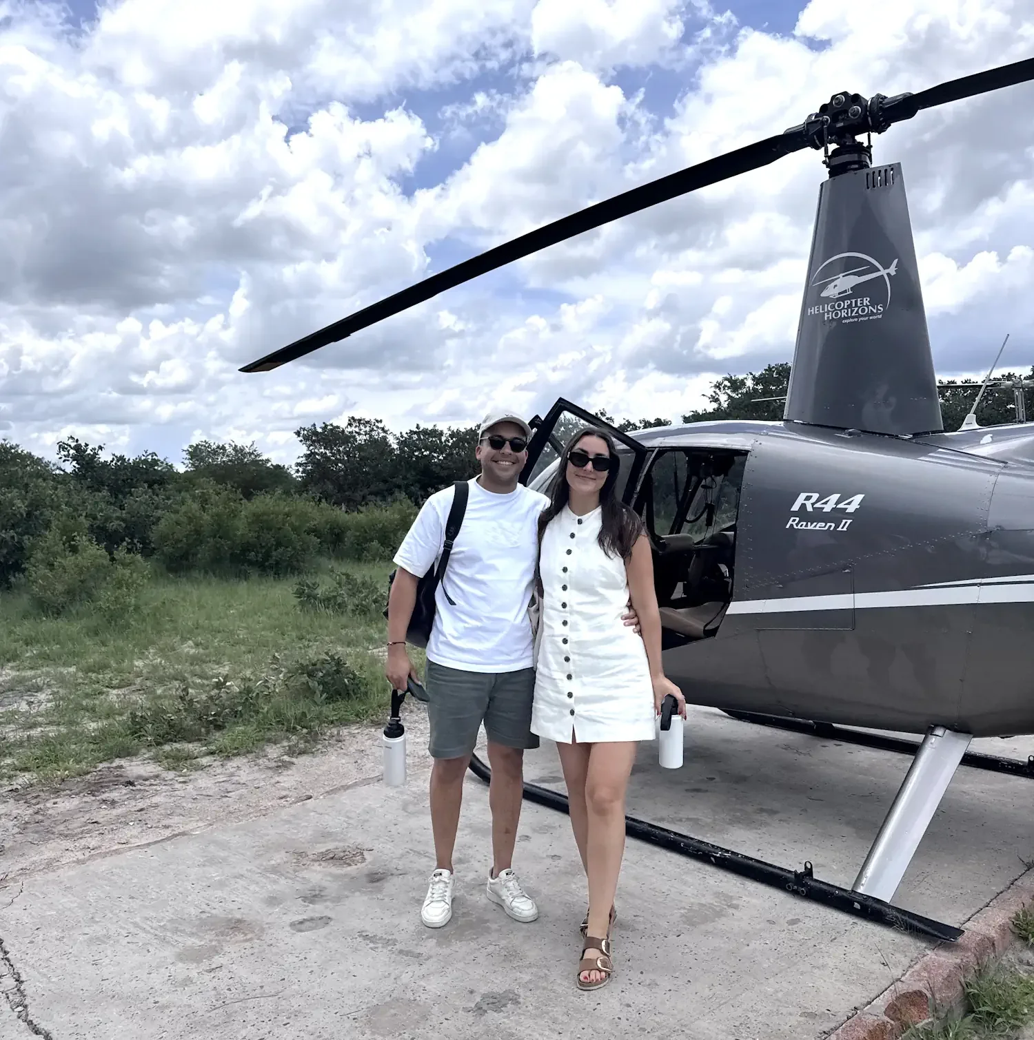 Co-founders James and Kristy Briscoe standing side-by-side and smiling in front of a parked, grey Helicopter Horizons safari helicopter on a bright day with a cloudy blue sky.