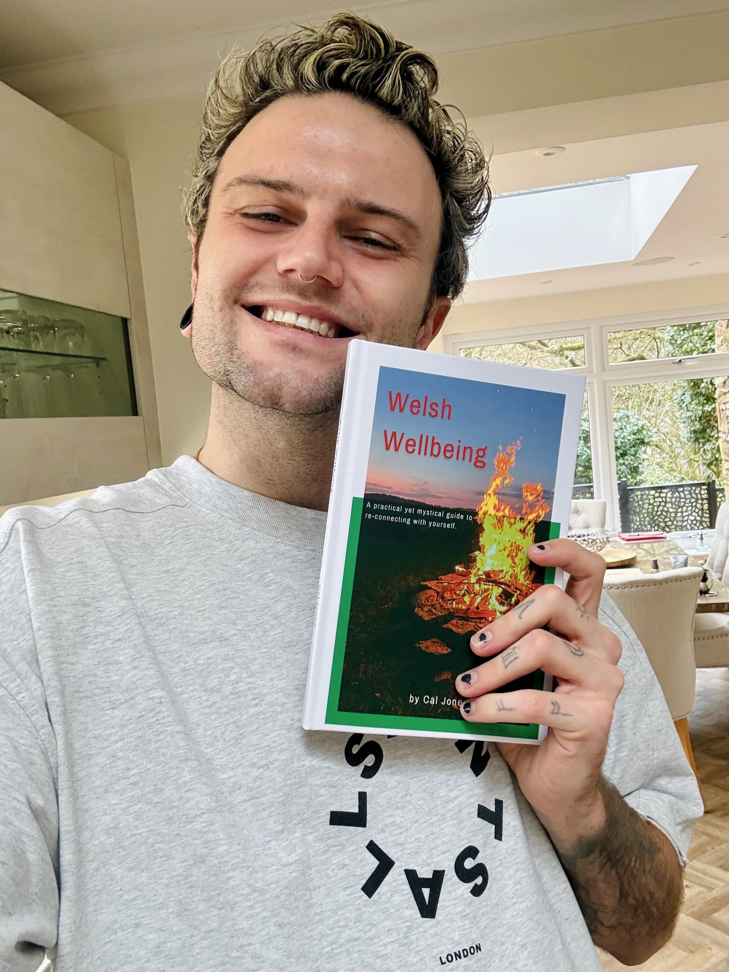 A smiling man with tattoos and a septum piercing holding a book titled 'Welsh Wellbeing' inside a well-lit room with large windows and greenery outside.