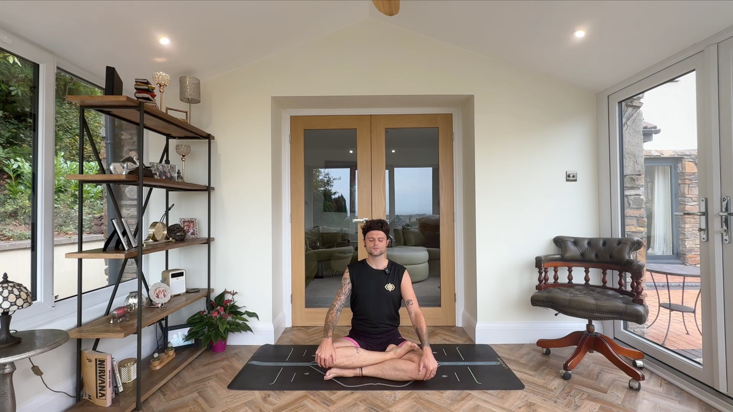 A man practicing yoga in a bright living room, sitting cross-legged on a yoga mat in a meditative pose with eyes closed, surrounded by modern decor and large windows showing an outdoor patio.