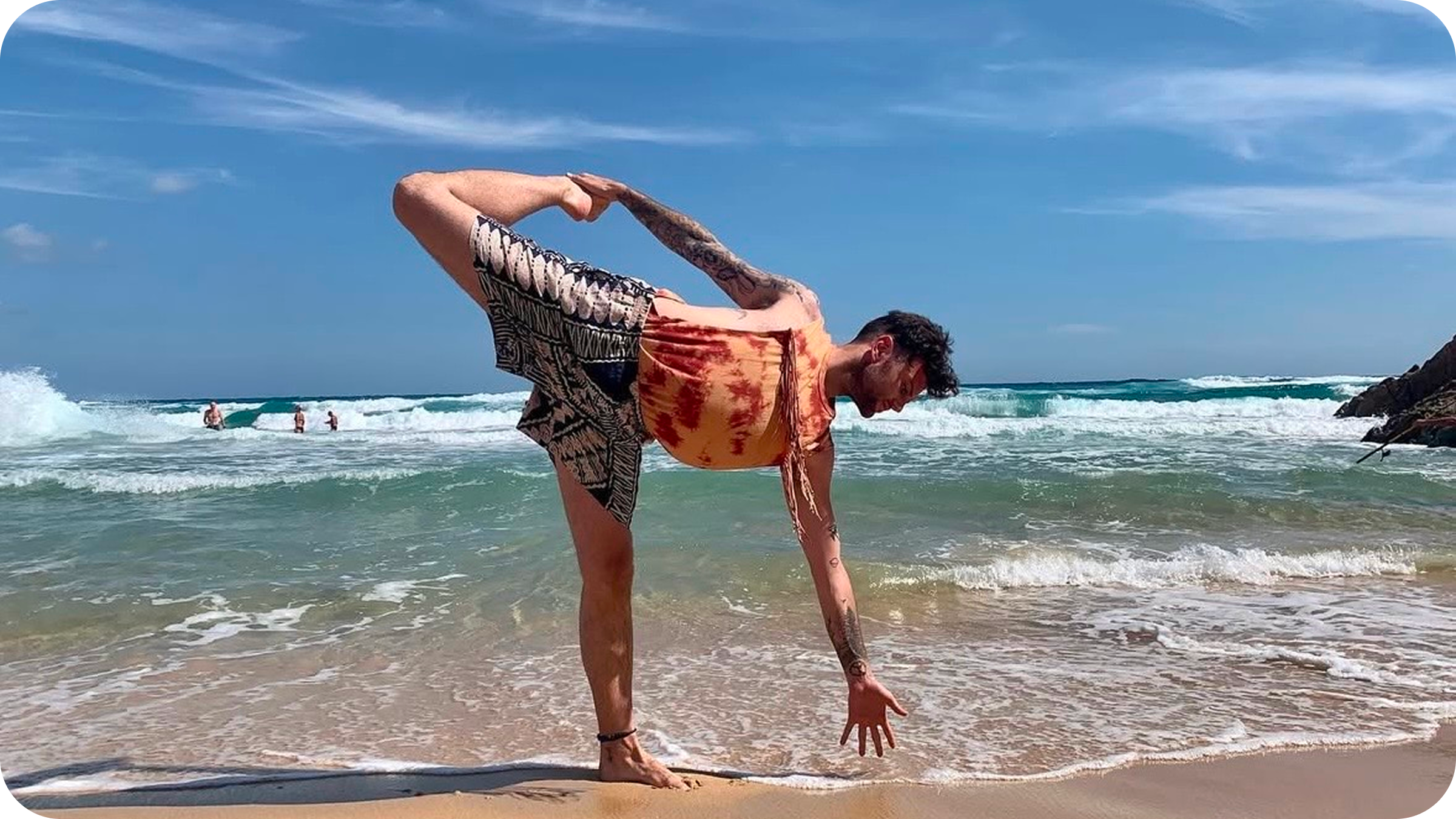 A man practicing yoga on a sandy beach near the ocean, balancing on one leg with his other leg raised behind him, holding his foot with his hand, during daytime with a clear blue sky.