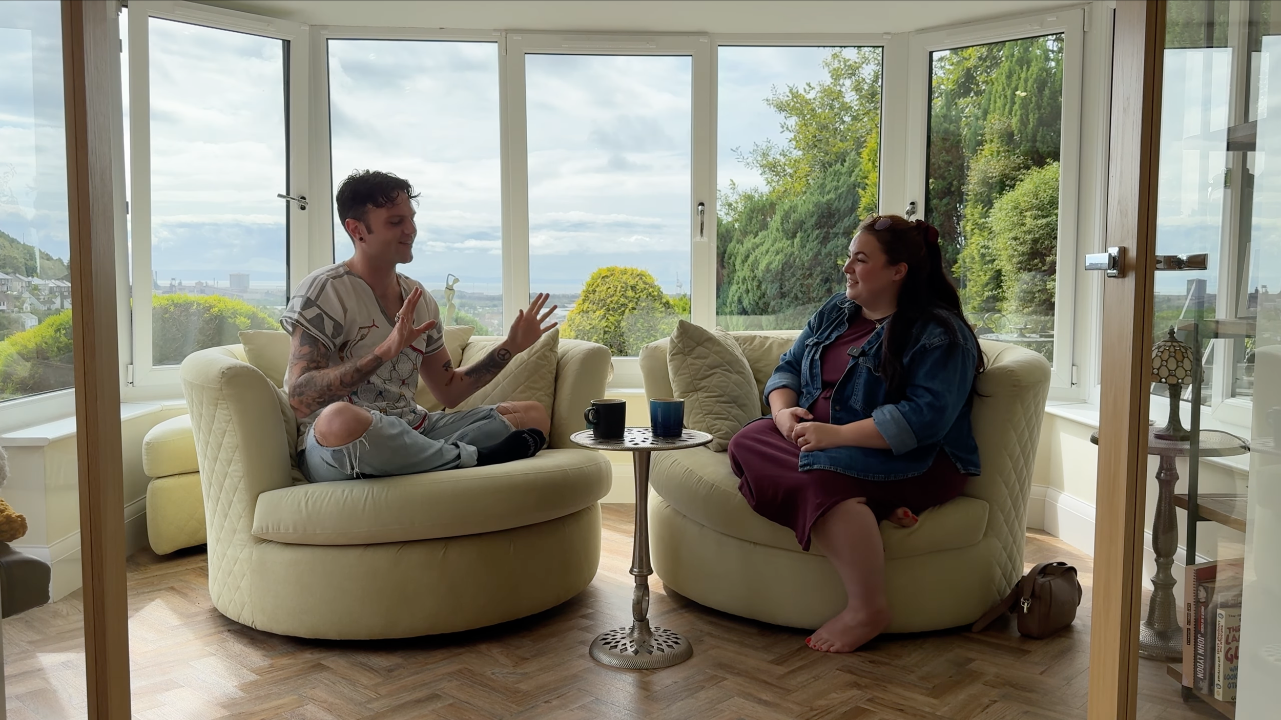 Two people sitting on yellow sofas in a bright living room with large windows and green trees outside, having a conversation. There is a small round table with cups between them.