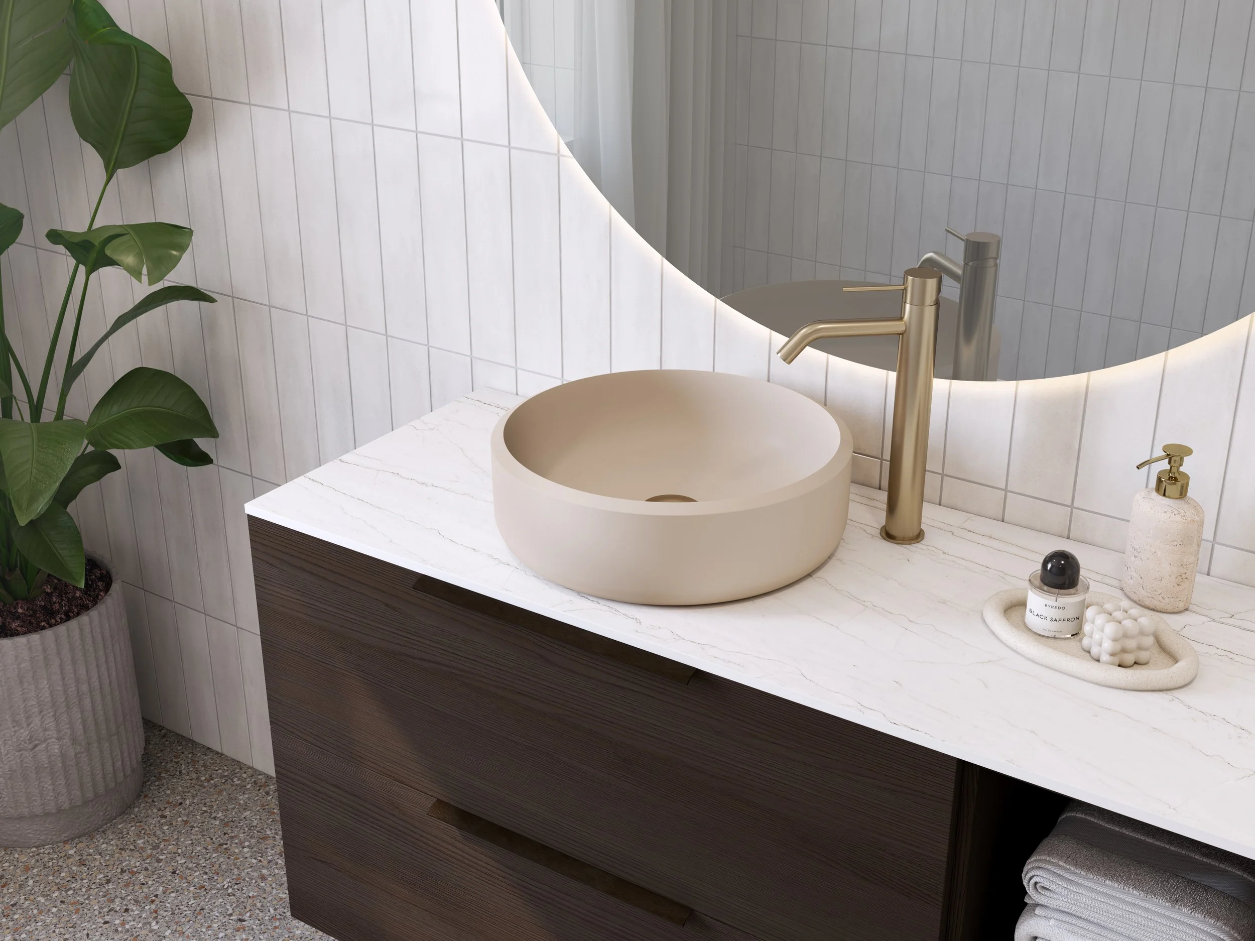 Badeværelsesvask i beige med guldfarvet armatur på hvid marmor bord, stor grøn plante i grå potte, væg med hvide fliser, spejl med integreret belysning, håndsæbe i pumpespand, parfume og badebold på oval skål, sammenrullede hvide håndklæder under bordet.