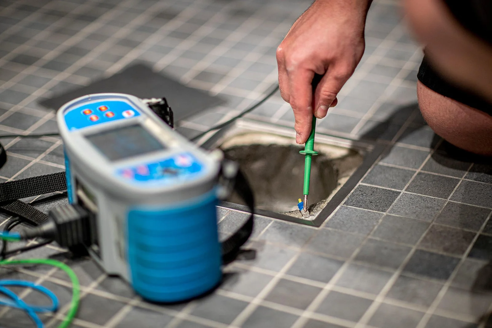 En person bruker et verktøy for å teste eller reparere en konstruksjon i gulvet, med måleutstyr ved siden av.
