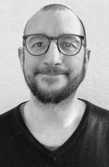 Et svart-hvitt portrett av en man med briller, skjegg og kort hår, smilende mot kameraet.