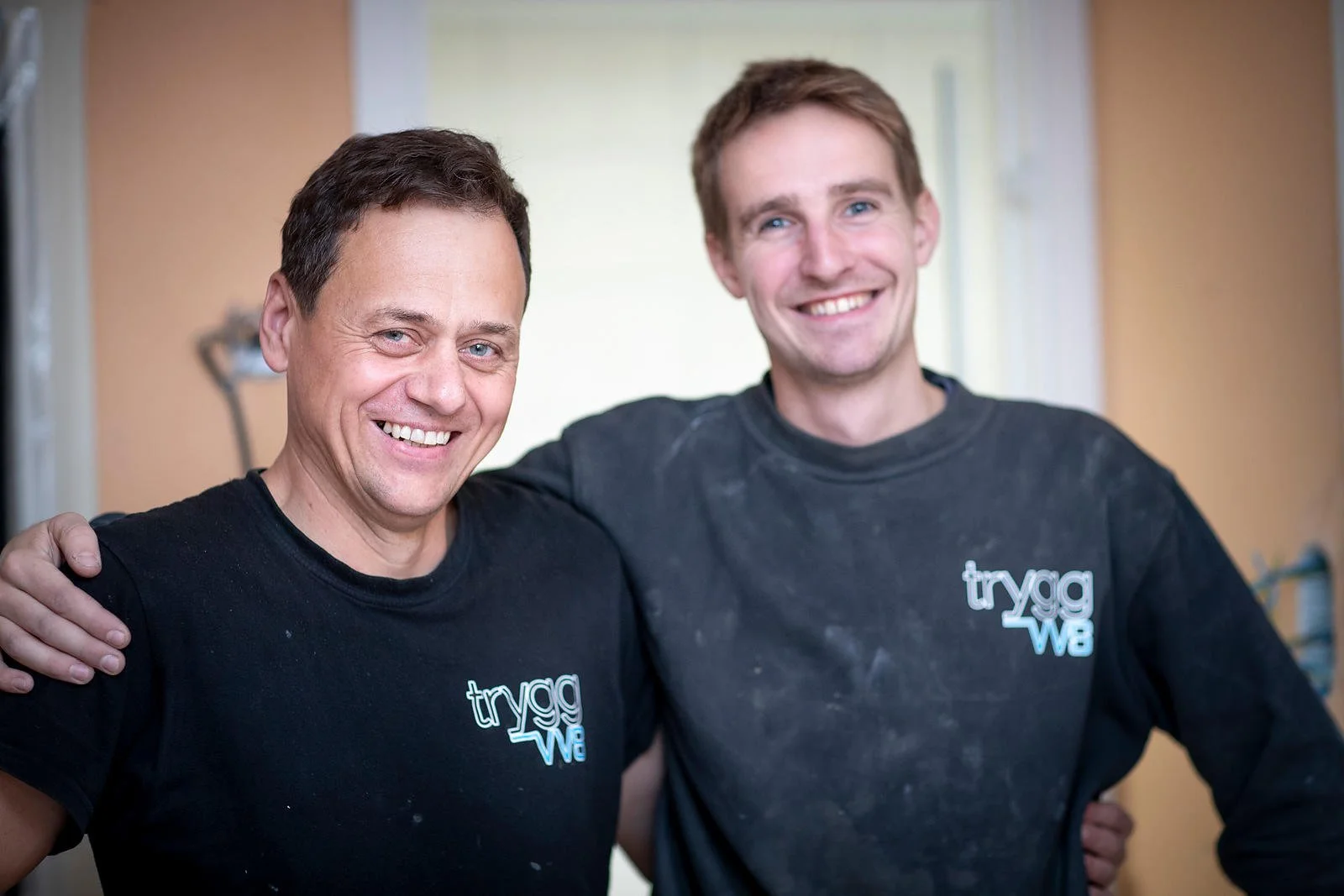 To menn smiler og har armer rundt hverandre, begge iført svarte tryggewe t-skjorter, inne i et rom med lyse vegger.