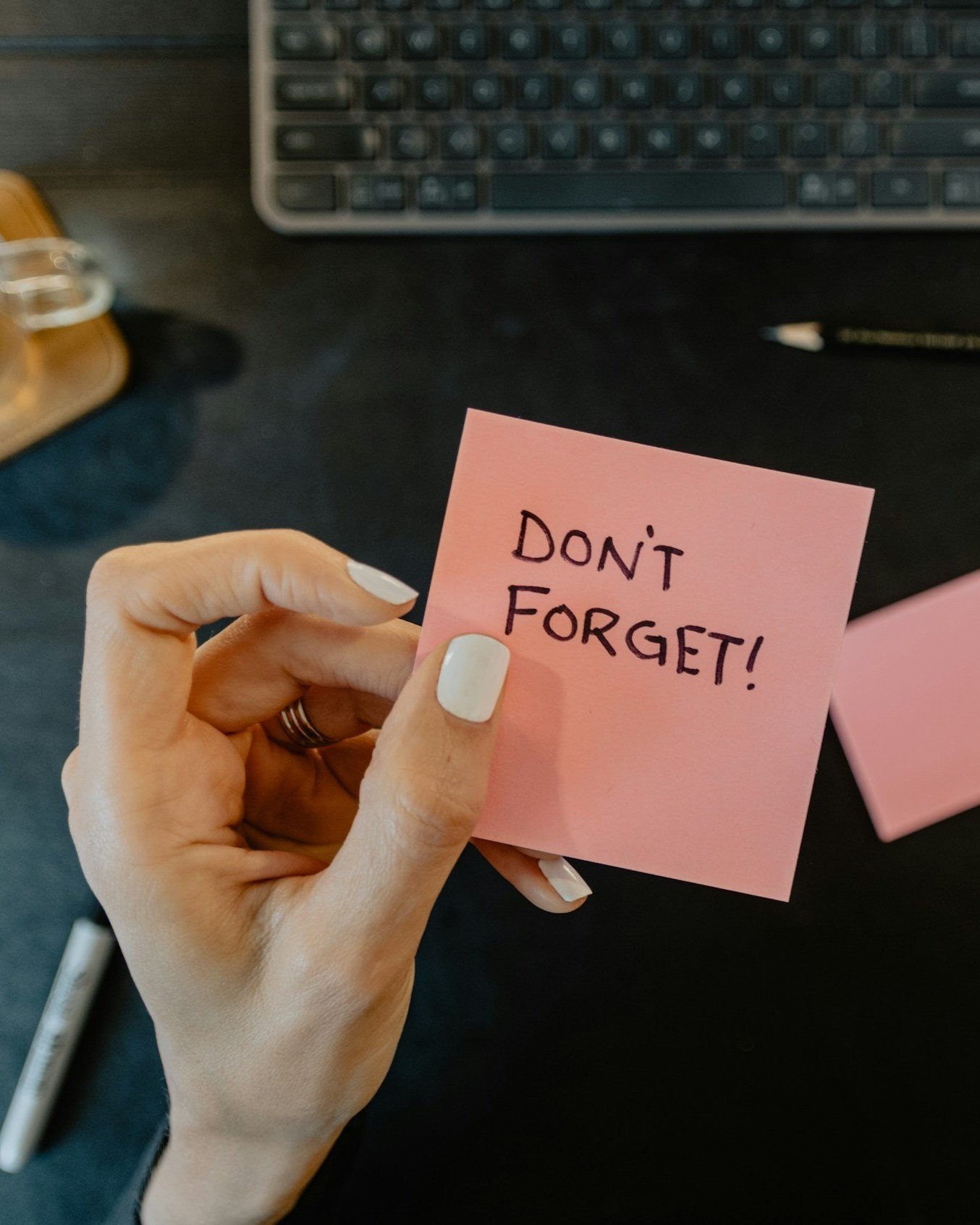 Hånd holder en rosa post-it lapp med teksten 'DON'T FORGET!' foran en datamaskin og et skrivebord.