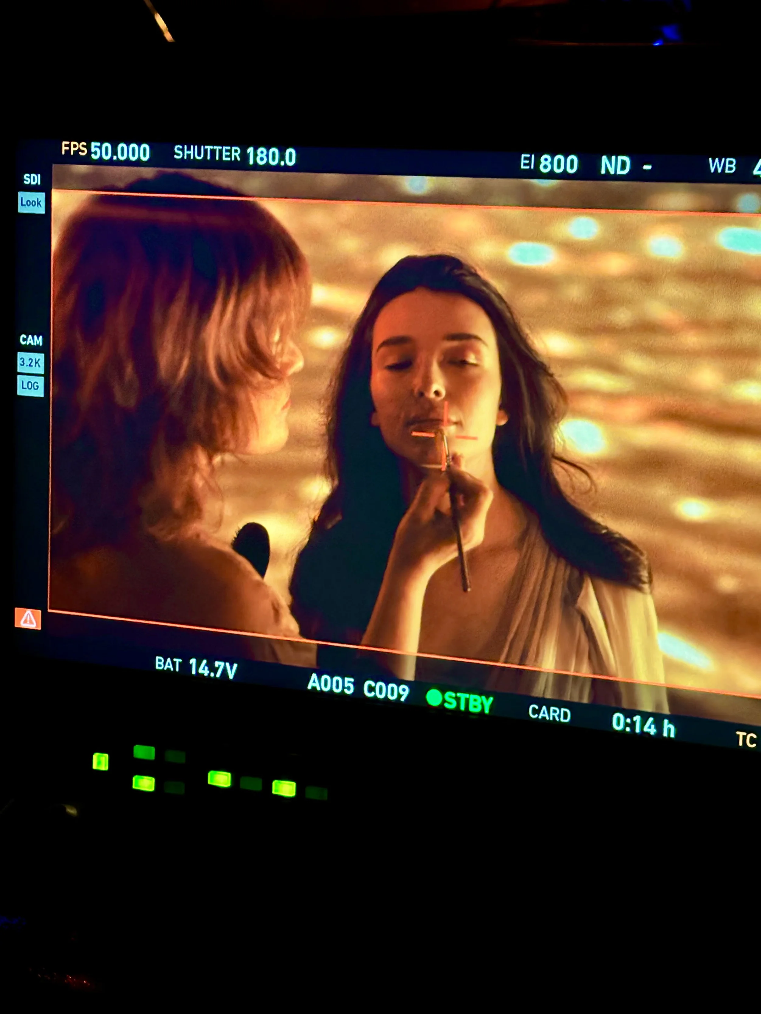 On a film set, a makeup artist applies lipstick to a woman with long dark hair, against a background of golden, bokeh-like lights.