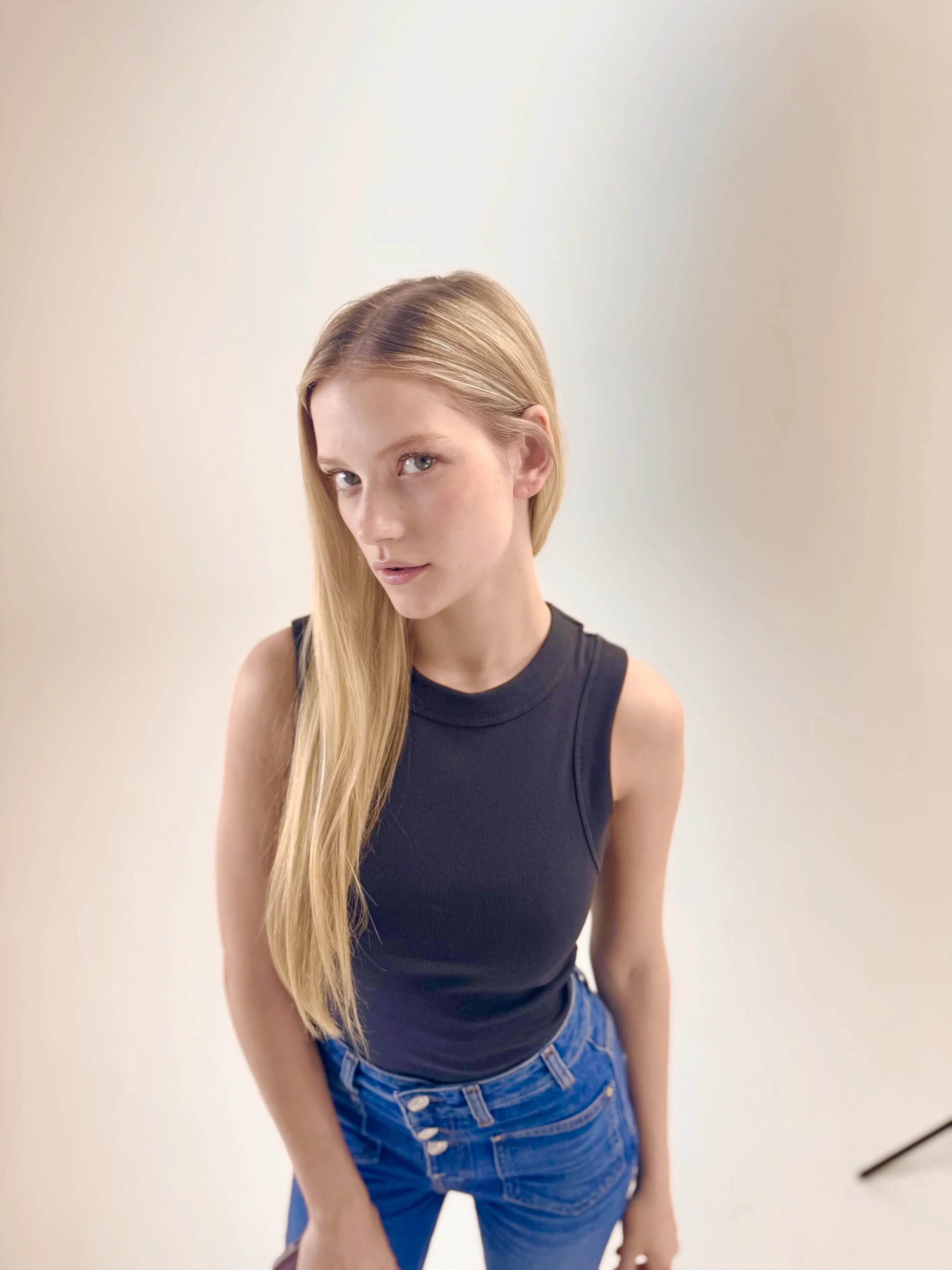 A young woman with long blonde hair, wearing a black sleeveless top and blue jeans, standing against a plain light-colored background.