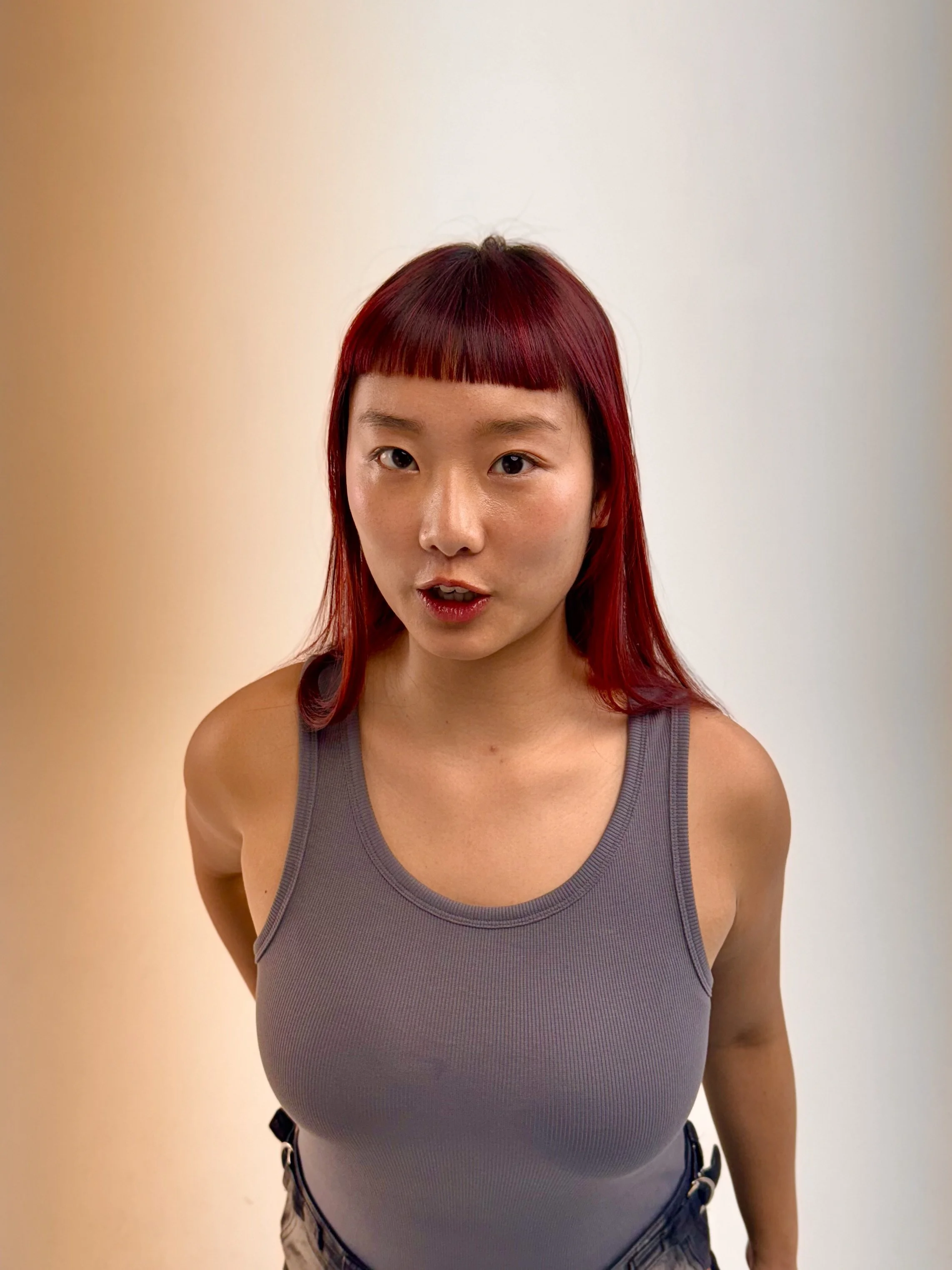 A young woman with red hair and bangs, wearing a gray tank top, standing against a plain background with warm colors on the left and cool colors on the right.