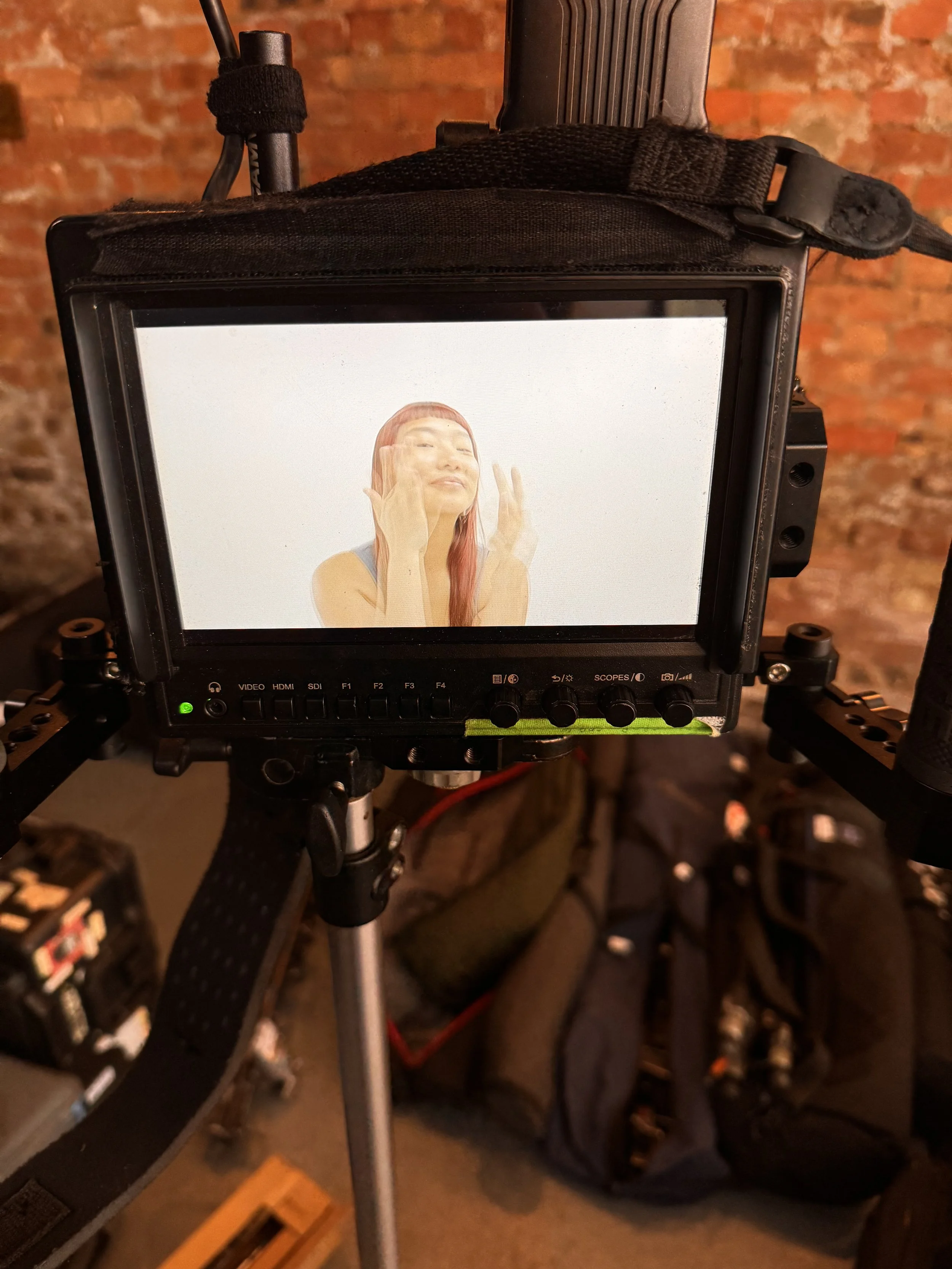 On a film set, a monitor displays a woman with reddish hair and a fringe, touching her face with her eyes closed against a plain background.