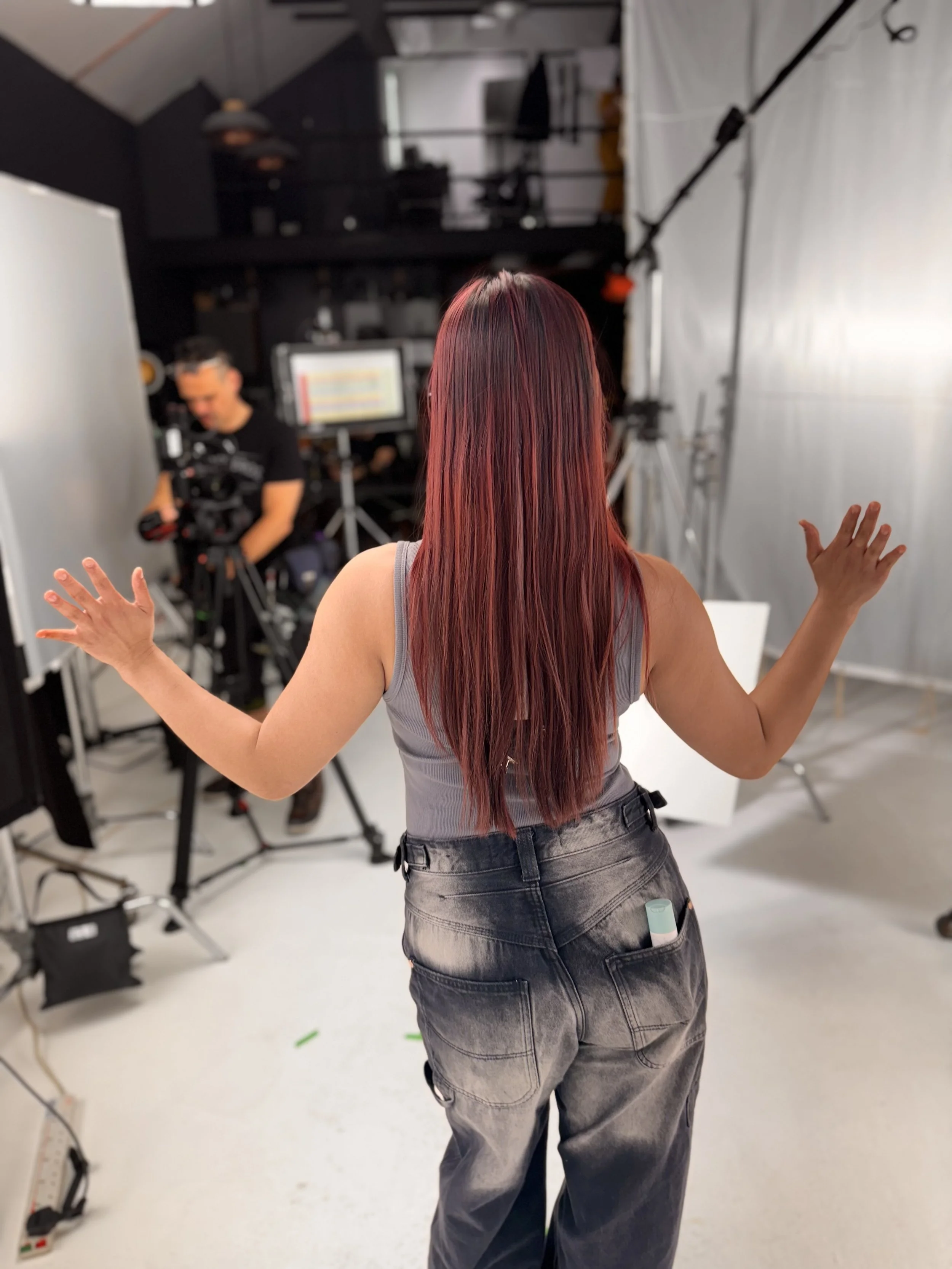 A woman with long red hair standing in a photography studio, facing away from the camera, with her arms slightly raised. Behind her, a man is adjusting camera equipment, and there are various studio lights and a monitor in the background.