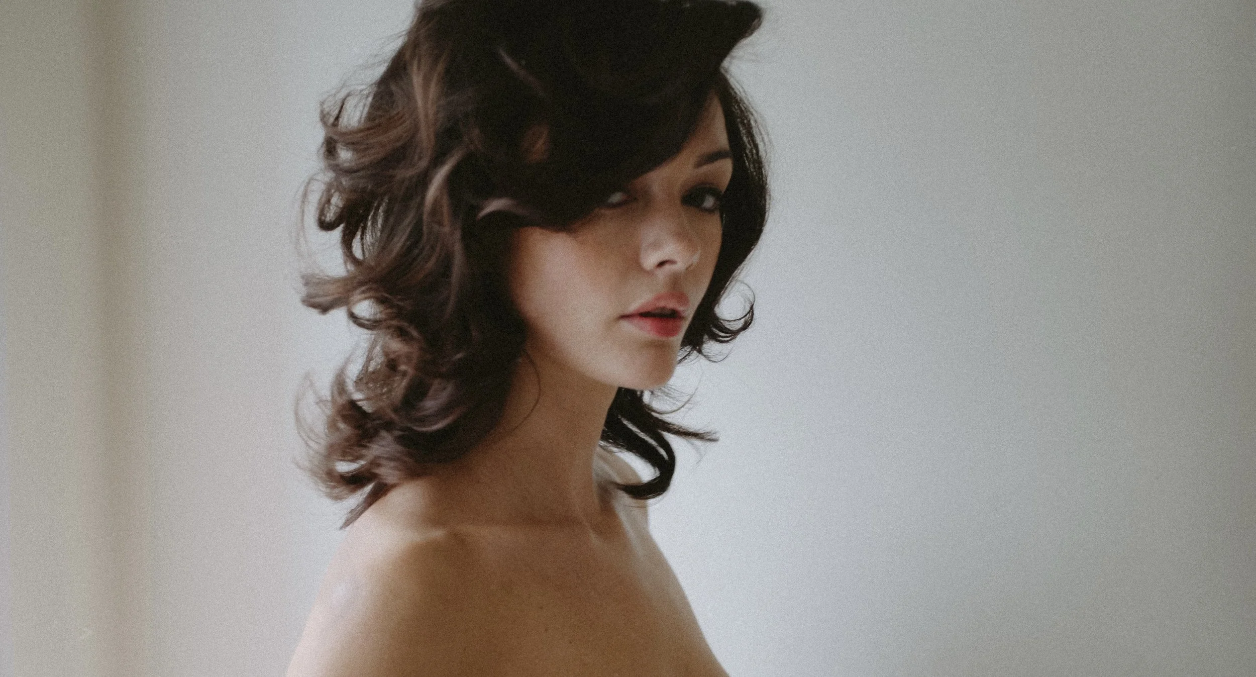 A woman with dark, wavy hair looking over her shoulder at the camera, with a neutral expression, against a plain light background.