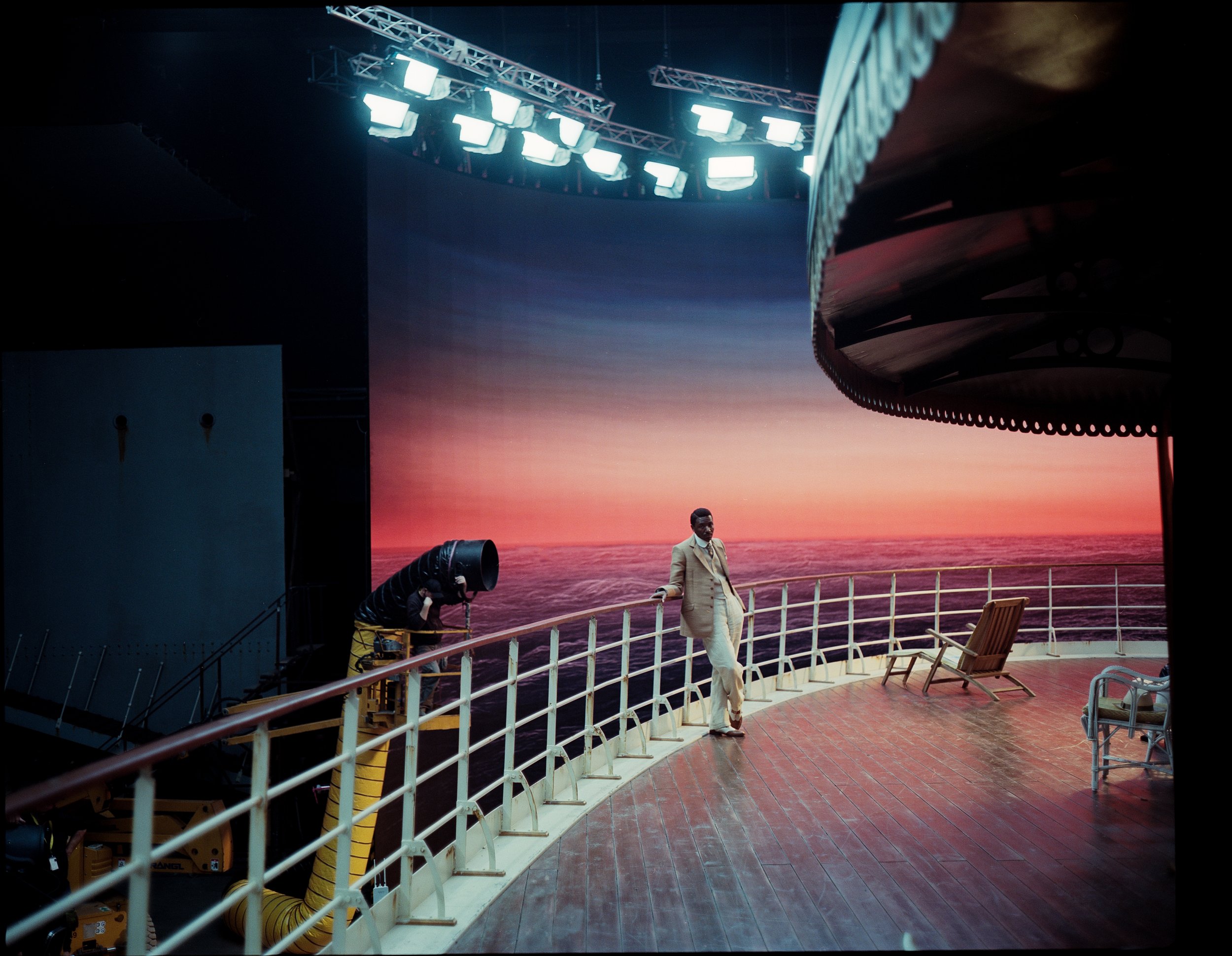 A man standing on the deck of a ship, with a sunset over the ocean background. The deck has lounge chairs and a railing, and there is a large screen or backdrop with sunset and ocean imagery. Bright lights are hanging overhead, indicating a film or p