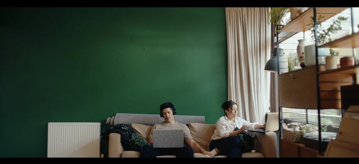 A man and woman sitting on a beige couch with a green wall behind them, each working on laptops. The woman has headphones on. There are large windows with beige curtains to the right, letting in natural light, and a wooden shelf with plants and decor