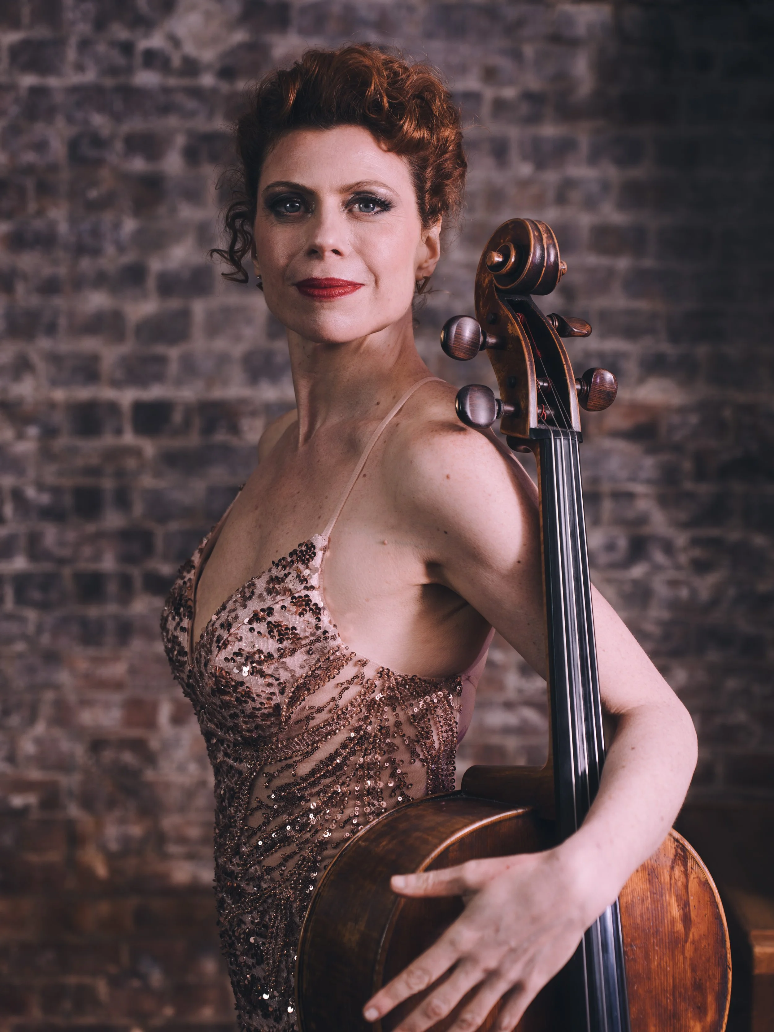 A woman with short curly red hair wearing a sparkly pink dress holding a cello against a brick wall background.