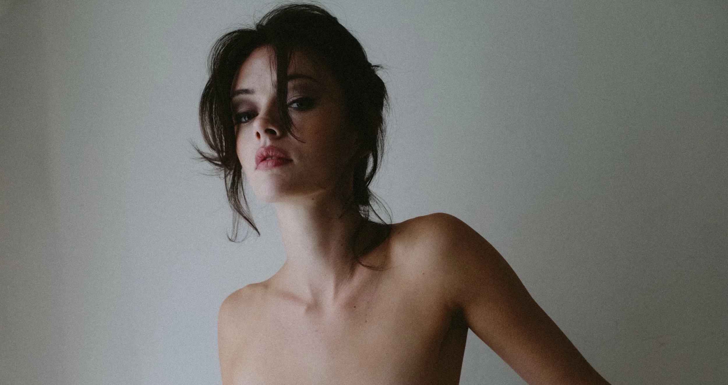 A woman with dark, tousled hair and minimal makeup poses topless against a plain light-colored wall.