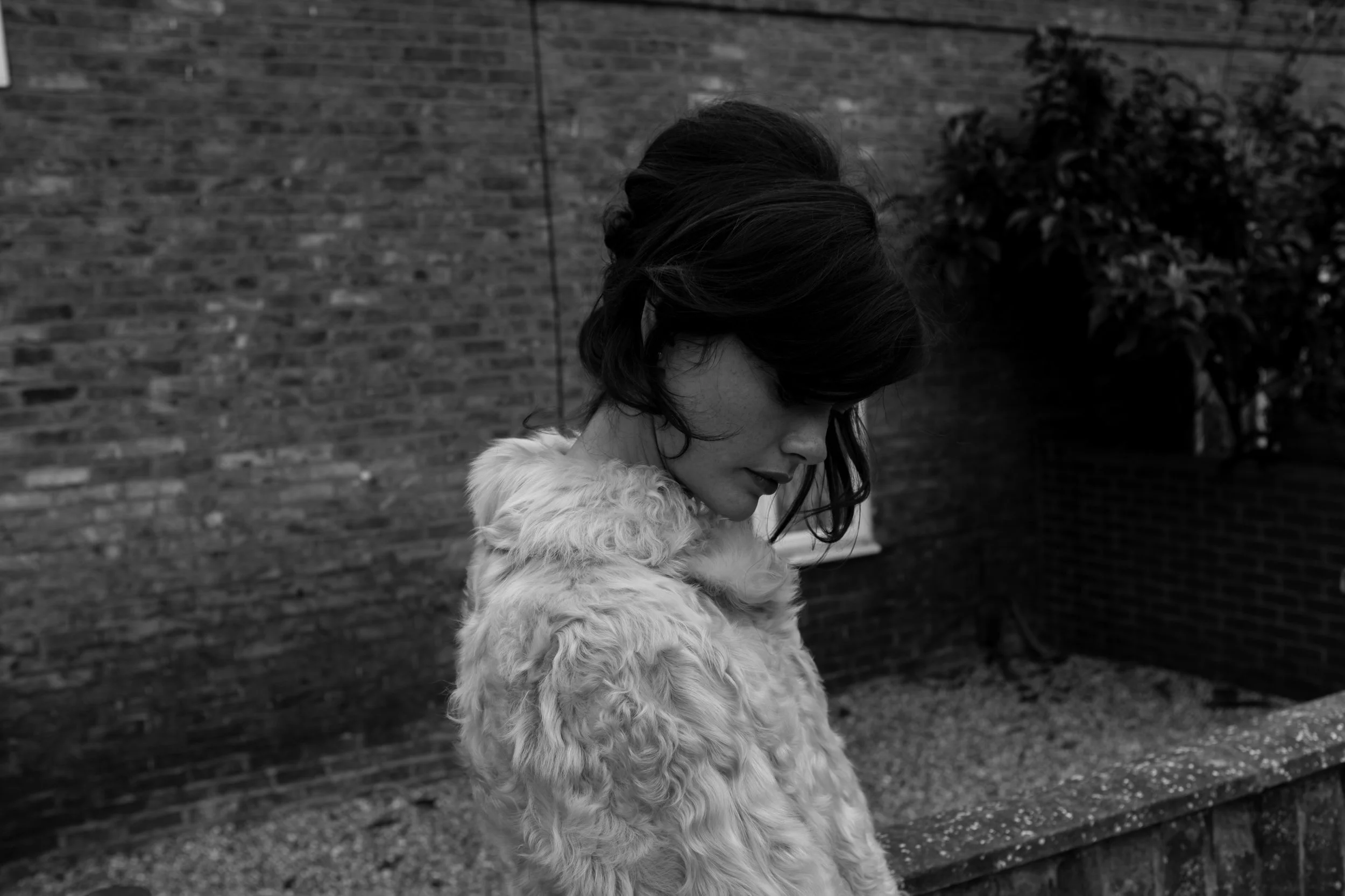 A woman with dark wavy hair looking downward, wearing a fluffy fur coat, standing outdoors near a brick wall and bushes in black and white.