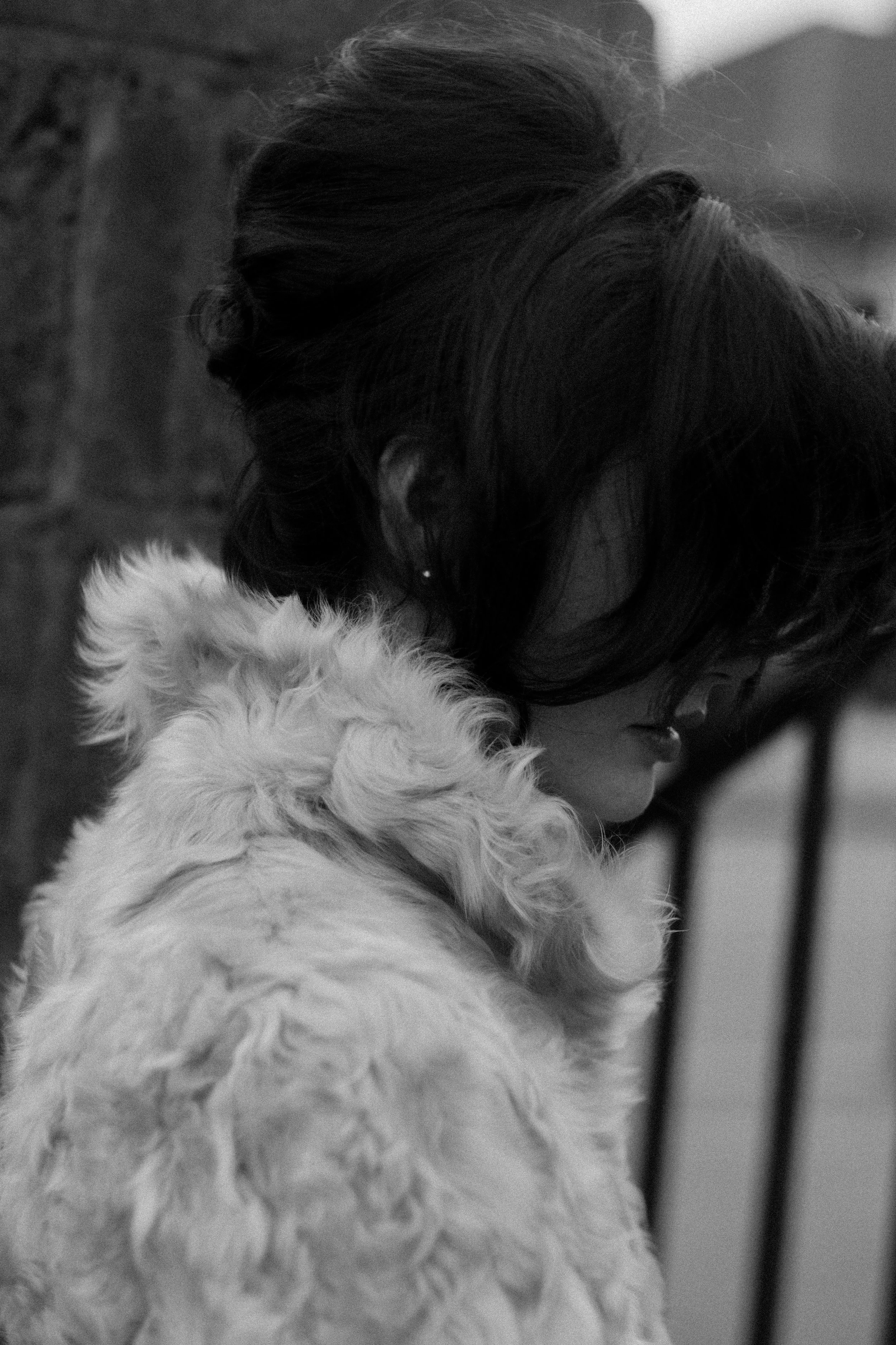 A woman with dark hair and glasses gently resting her head and cheek on a fluffy, light-colored dog, both facing downward.