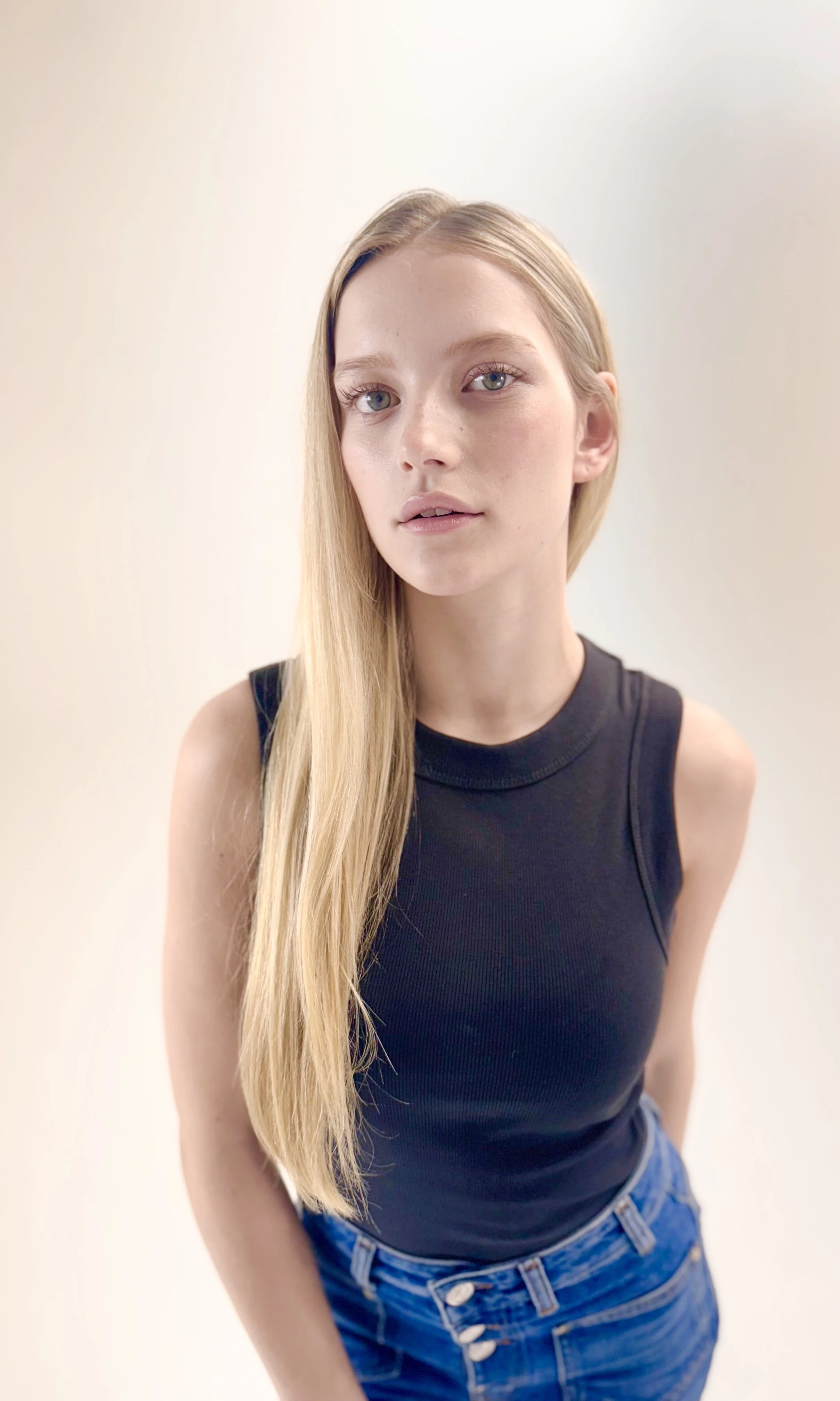 A young woman with long blonde hair, wearing a black sleeveless top and blue jeans, standing against a plain light-colored background.