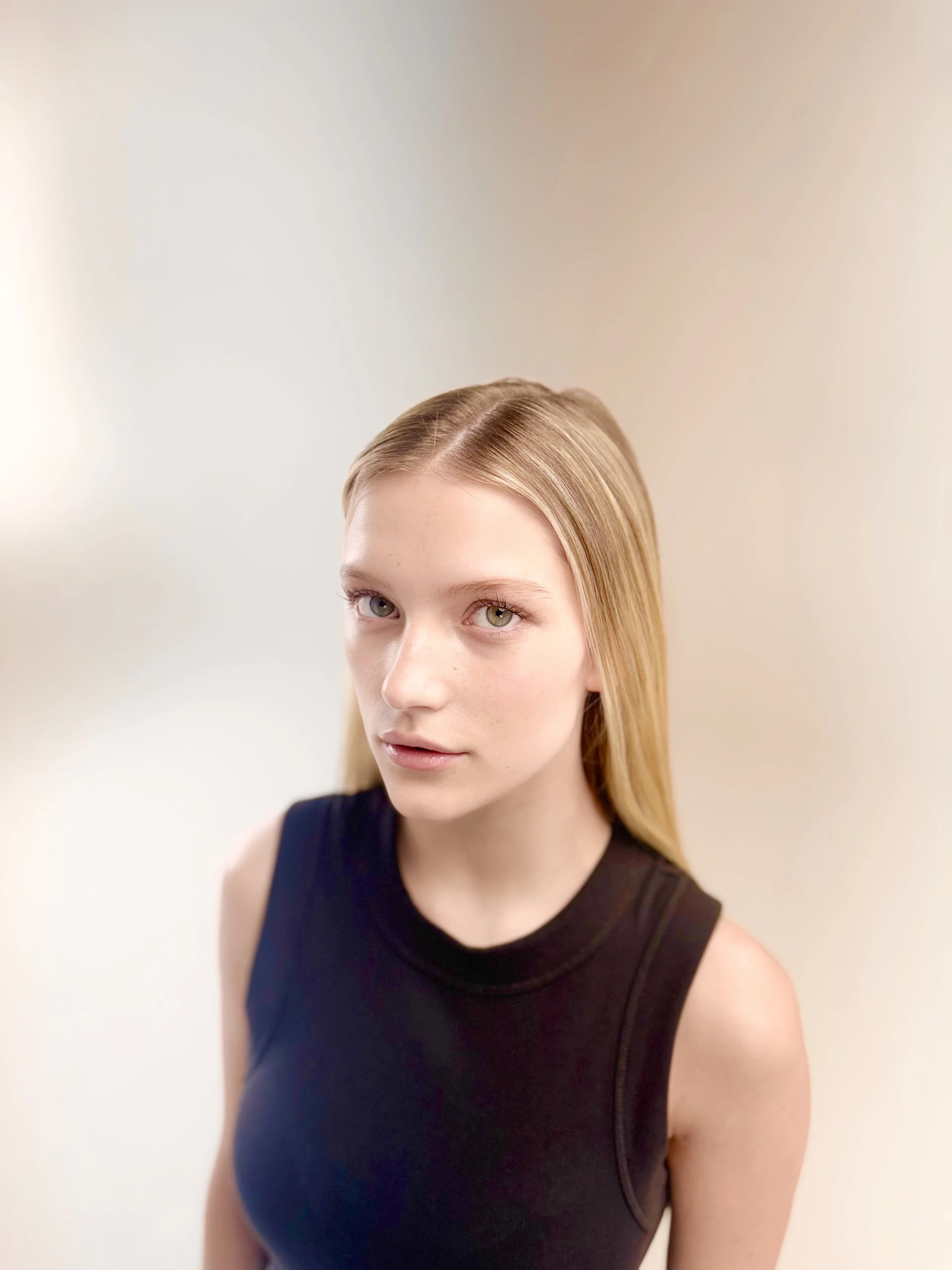 Portrait of a young woman with blonde hair wearing a sleeveless black top, standing against a plain light-colored background.