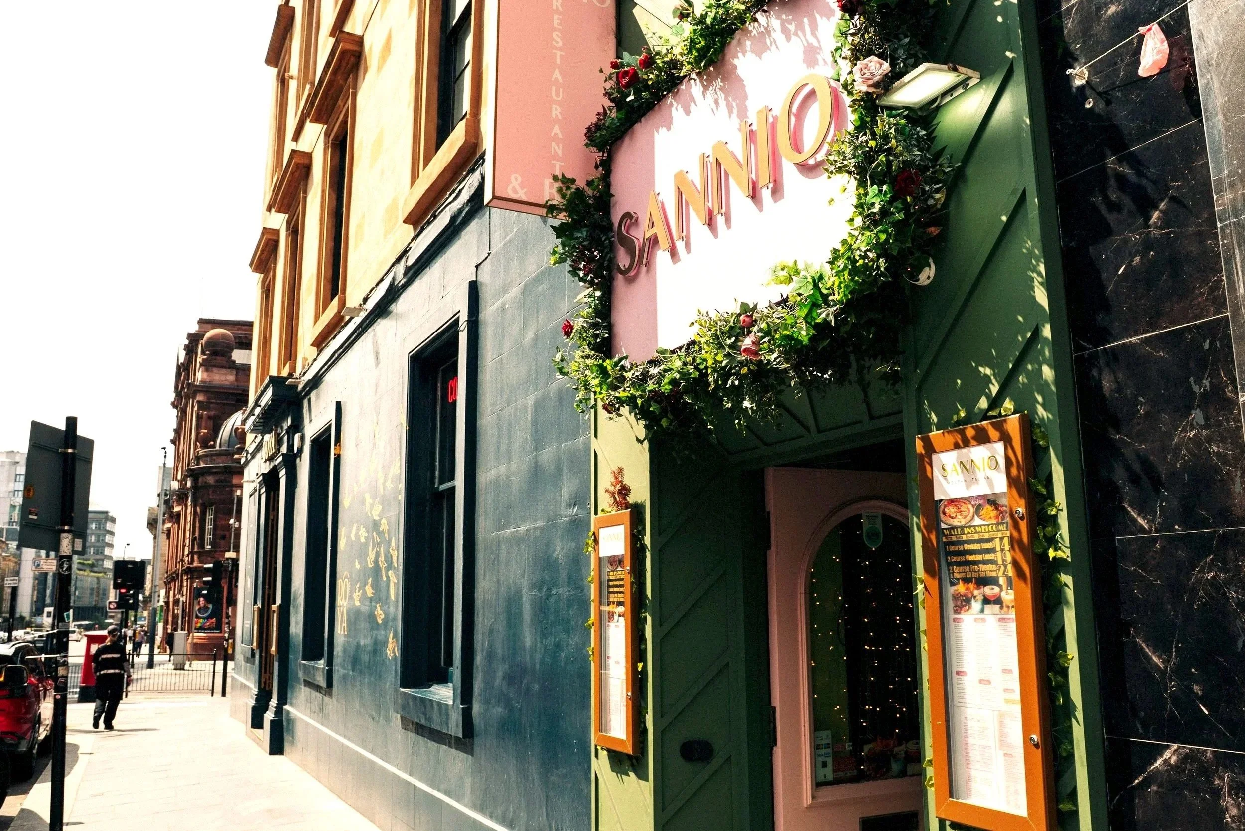Street view of a restaurant named SANNIO with a green facade, pink sign, and floral decorations, located on a city sidewalk with pedestrians and cars.