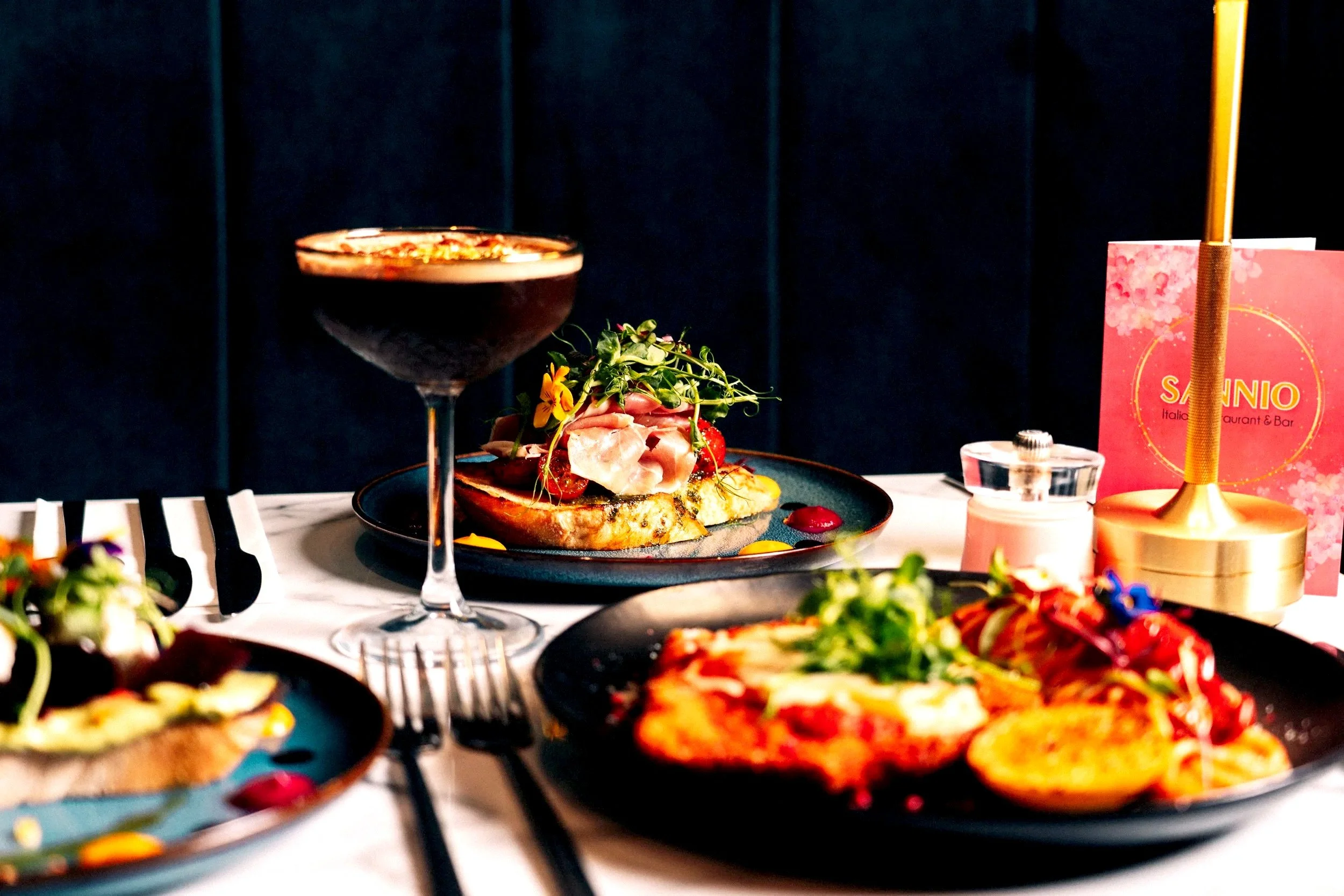 A table with various dishes, including a dark cocktail, a plate of gourmet food topped with microgreens and edible flowers, another plate with pasta, and a slice of bread with toppings. There is a pink menu card, salt shaker, and golden candle holder in the background.