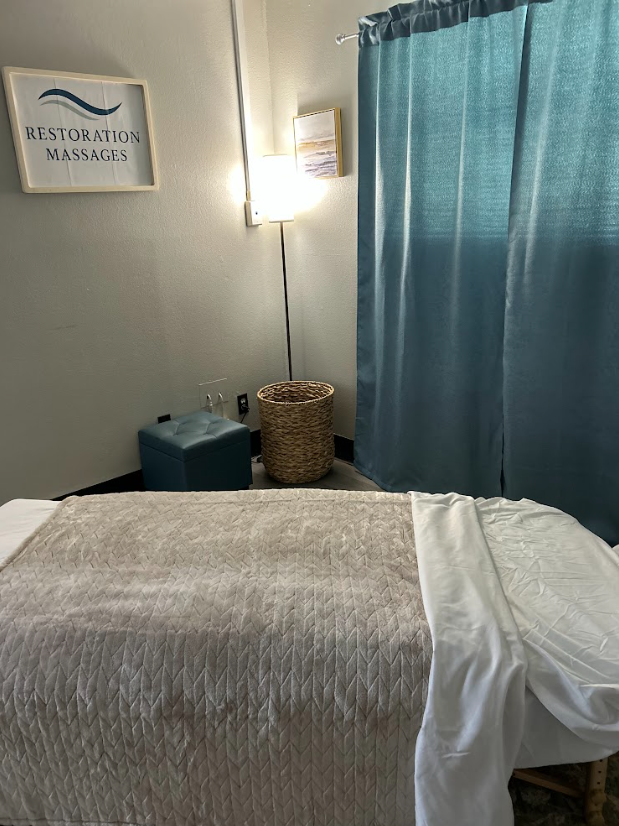 Spa room with massage table, blue curtains, a framed sign reading 'Restoration Massages,' a floor lamp, a woven basket, and a small storage ottoman.