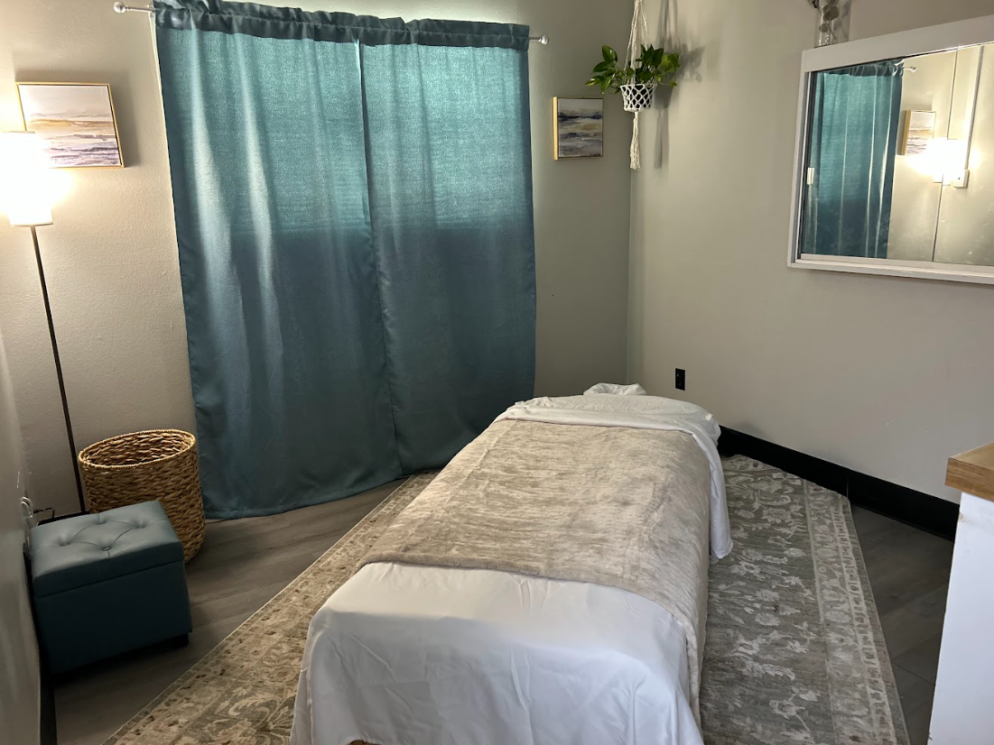 A massage room with a bed, blue curtains, wall art, a standing lamp, a wicker basket, an upholstered stool, and a hanging plant next to a mirror.