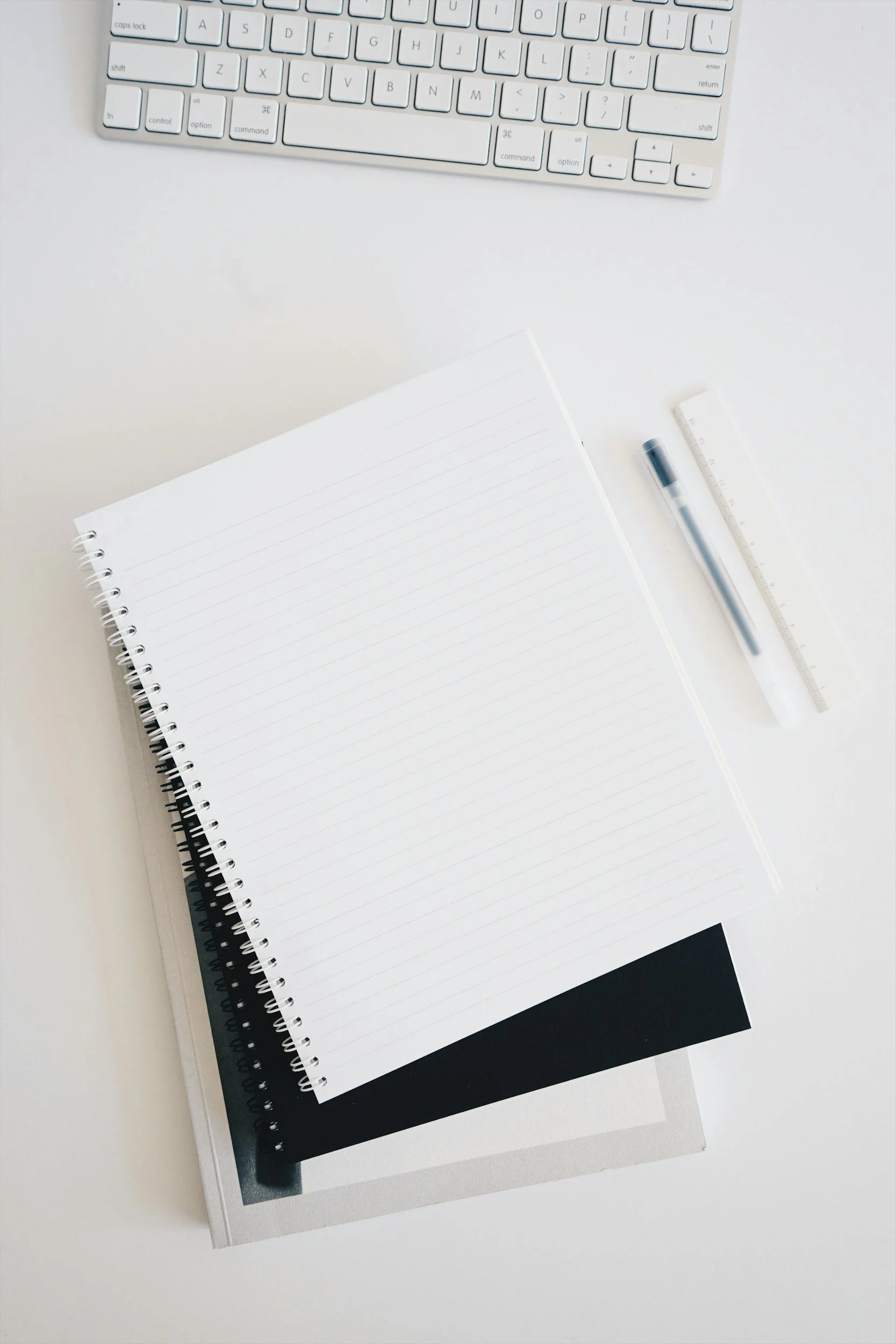 A white computer keyboard, a stack of three notebooks or sketchbooks with spiral and hardcover bindings, a clear ruler, and a black pen on a white desk.
