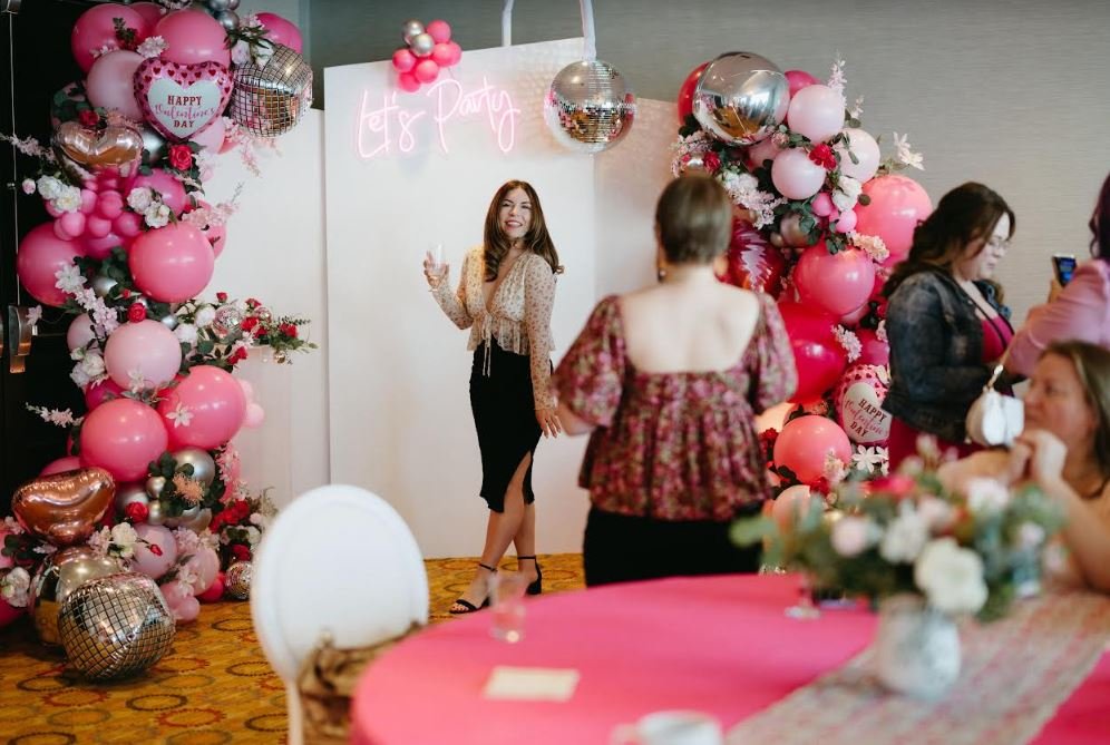 Group of people at a party with pink and red balloon decorations, floral arrangements, and a 'Let's Party' neon sign. One person poses for a photo in front of the decorations, while others socialize and take pictures. The setting suggests a festive c