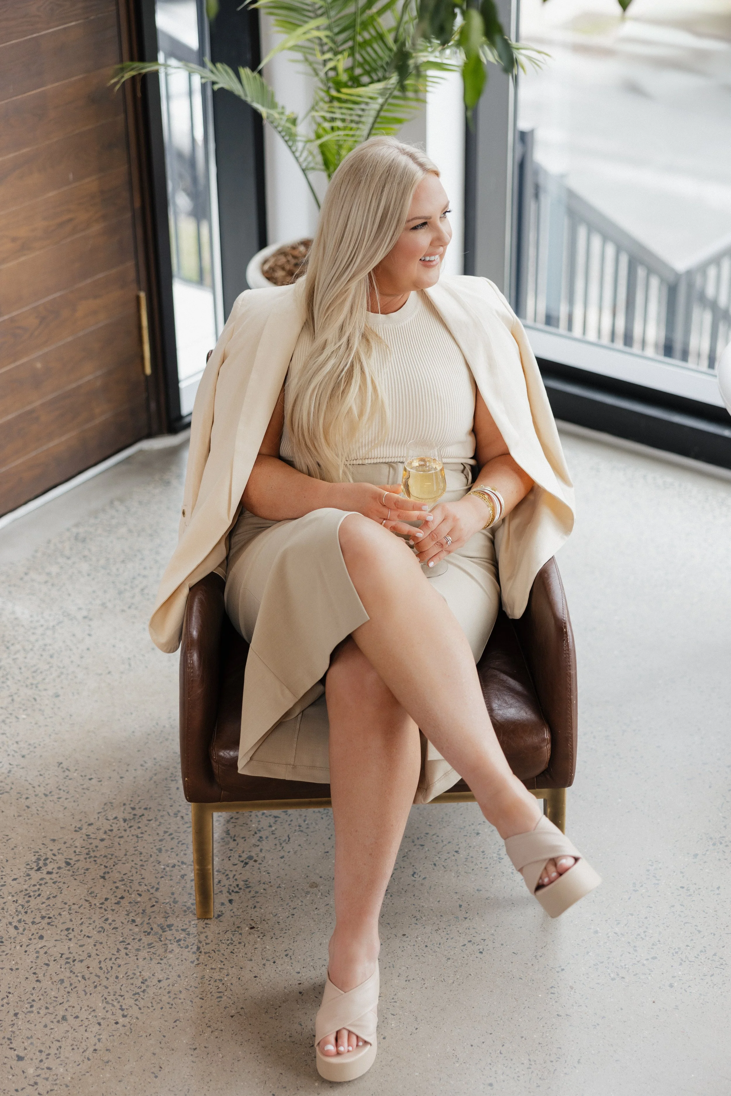 Woman sitting in a chair holding a drink, wearing beige outfit, long blonde hair, plant in background.
