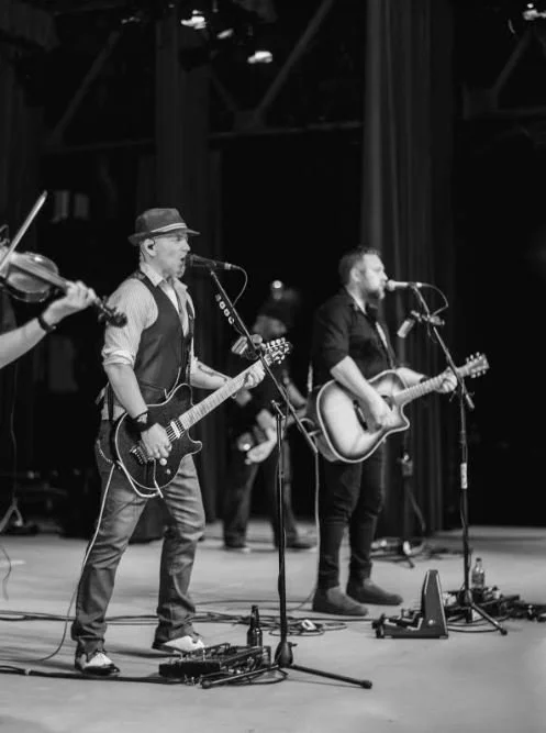 Black and white photo of a band performing on stage with guitars and a violin. Two musicians are singing into microphones.