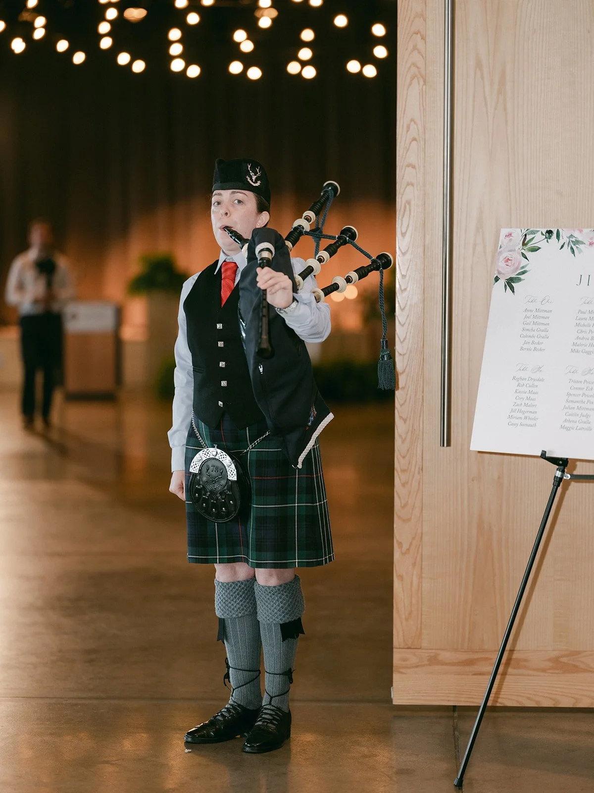 Person in traditional Scottish attire playing bagpipes indoors with a menu or signboard on the side.