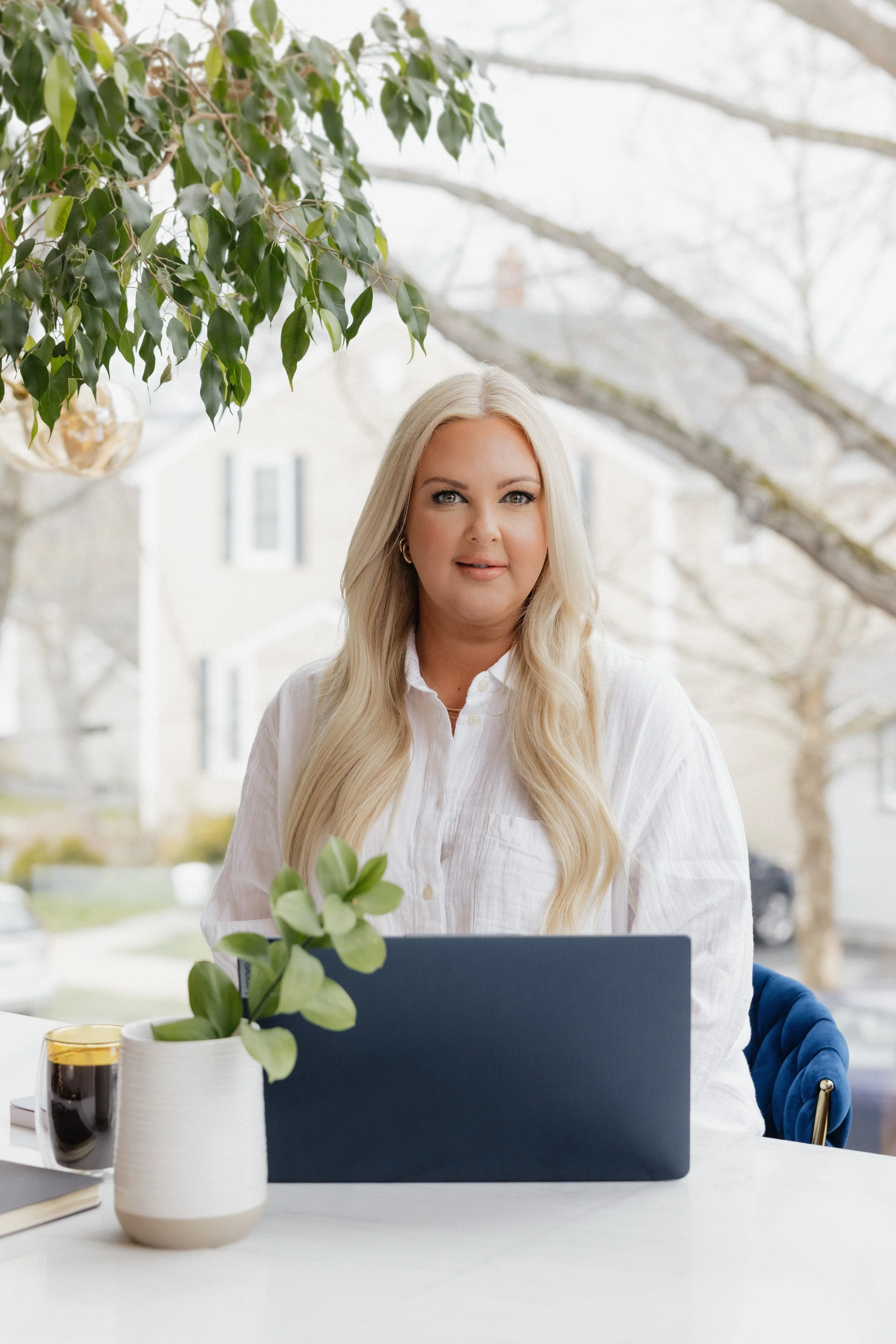 Woman with long blonde hair, wearing a white blouse, seated at a table with a laptop, plant, and coffee, with a tree and suburban neighborhood visible through a window.