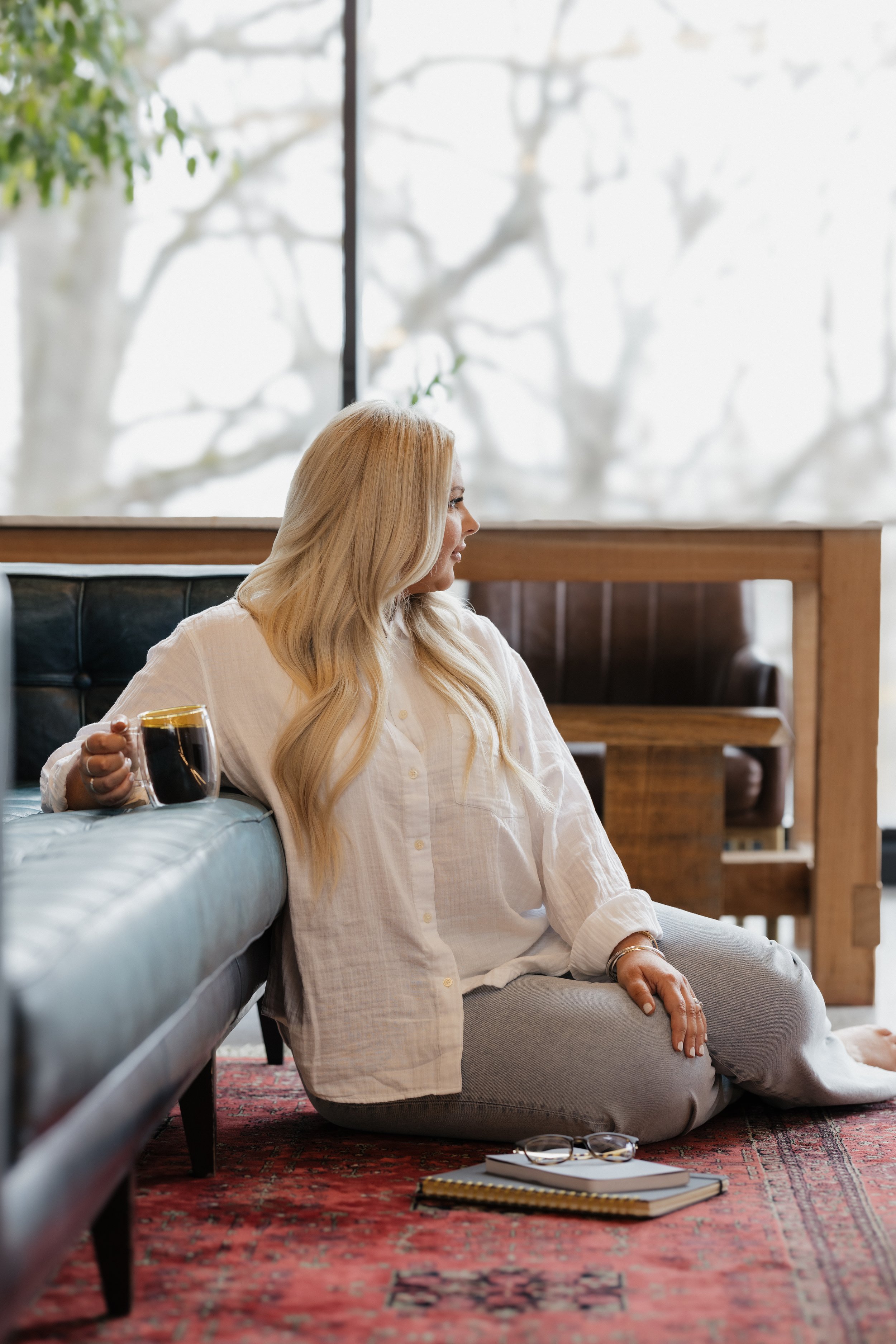 Woman sitting on floor next to couch with coffee cup, looking out window. Notebook and glasses on rug.