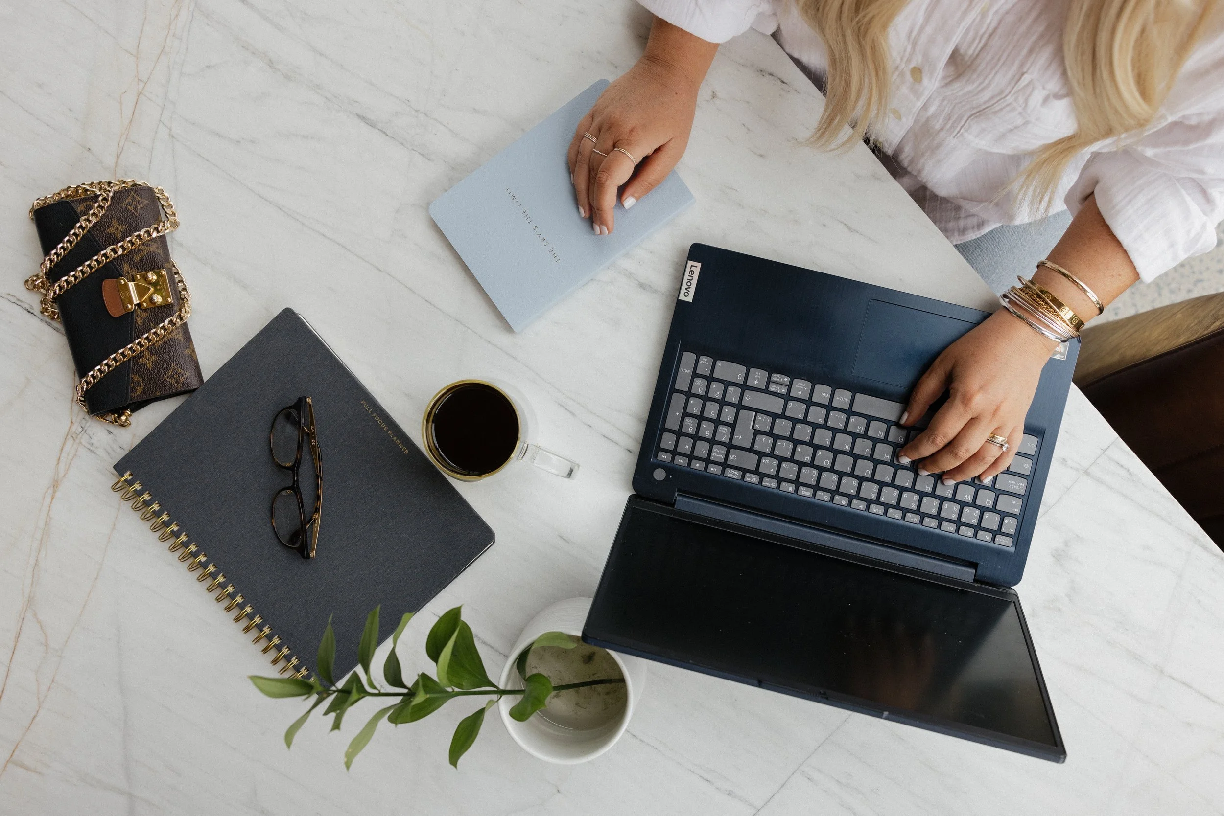 Woman working at a marble desk with a laptop, notebook, coffee, and a plant.
